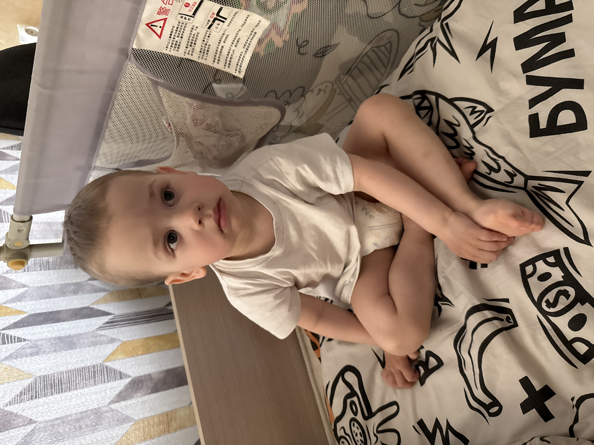 A young boy with light hair and light eyes is captured sitting upright in a playpen or crib in Rusanovo, Russia. He is wearing a white t-shirt and a diaper, with his legs bent and hands near his feet, looking directly at the camera with a neutral and curious expression.
The playpen features grey mesh sides and a grey frame. A white warning label with Chinese text is affixed to the mesh, displaying a red warning triangle and characters such as "警告" (Warning), "可见缝隙" (Visible gaps), and "床空服" (Bed empty clothing/bedding), alongside other instructional text regarding child safety and proper use of the crib.
The child is seated on a white blanket or sheet inside the playpen, which is decorated with bold black graphic patterns. These patterns include a stylized fish, lightning bolts, money symbols (dollar signs), and large letters, notably Russian text that reads "БУМА" (BUMA).
Beyond the playpen to the left, a wall is visible, featuring a geometric pattern composed of interlocking shapes in shades of white, grey, and yellow-gold. The scene is well-lit, indicating daytime, and set indoors, likely within a child's room or play area. The child's activity is primarily sitting and observing.