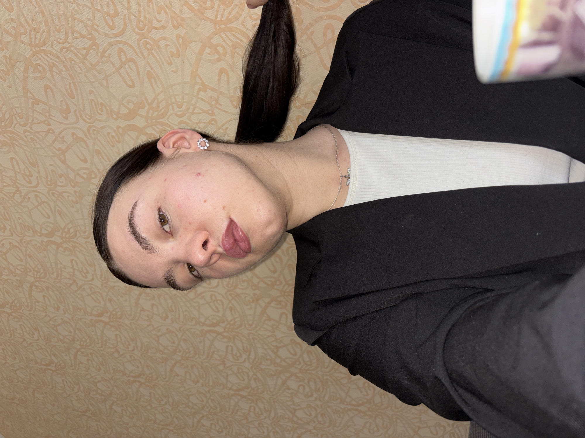 A person is posing for a selfie in what appears to be an interior setting with patterned wallpaper. The individual has dark hair, pulled back, with a section held up. They are wearing a black blazer over a light-colored top and accessorized with a small circular earring and a delicate necklace. The location is potentially the city of Perm, Russia, but without additional context or information within the image, this cannot be verified forensically.