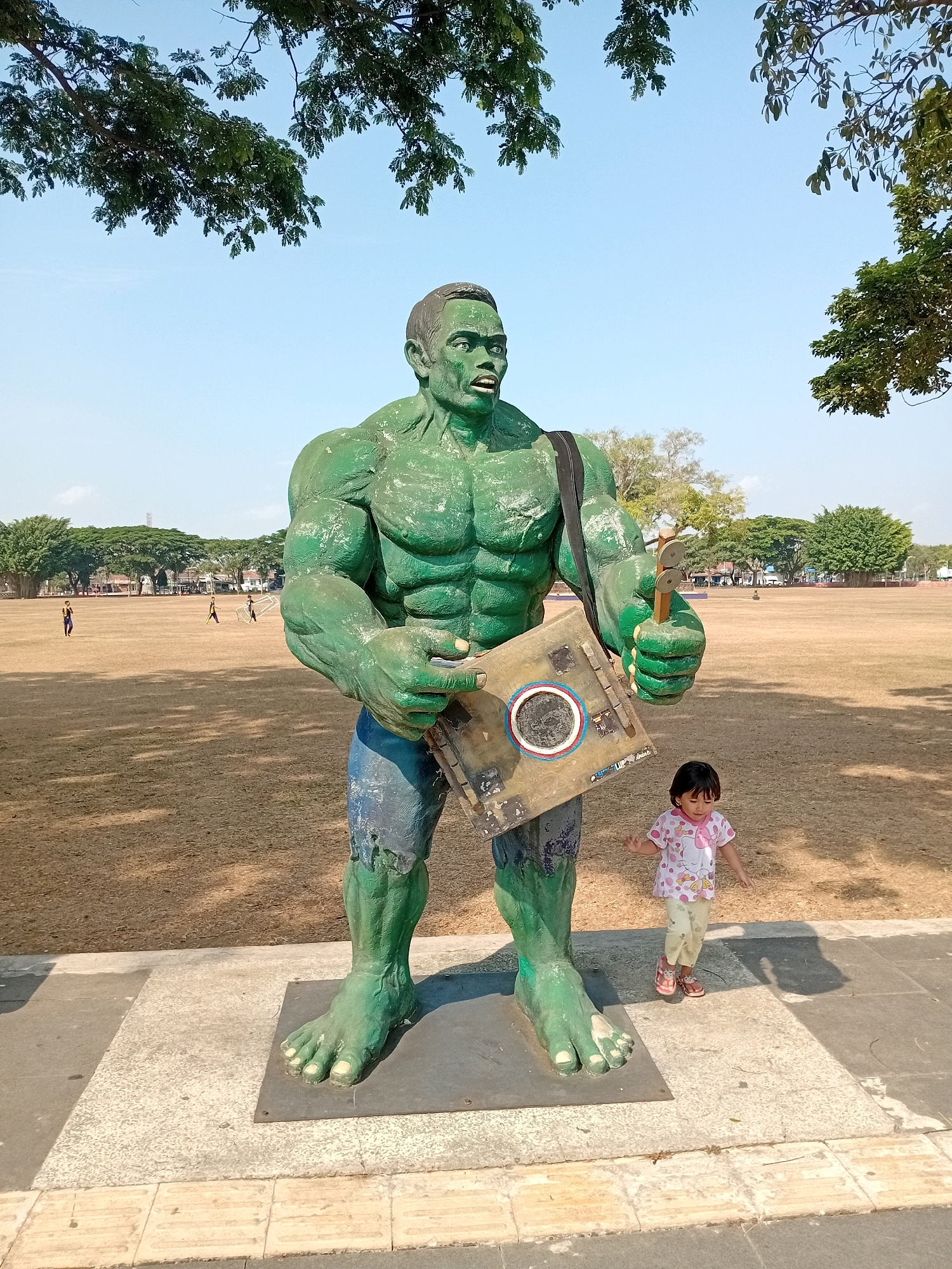 In Purworejo, Indonesia, a large, green statue of the Hulk stands in a park-like setting. The statue is wearing tattered blue shorts and has a black strap over its shoulder, from which hangs a makeshift drum or target. The Hulk is holding what appears to be a wooden percussion instrument. To the right of the statue, a young girl in a pink and white patterned shirt and light-colored pants, wearing sandals, is captured mid-movement, her arms extended. The ground in front of the statue is paved with textured tiles, leading to a large, dry grassy field. In the background, several trees line the edge of the field, with a clear blue sky overhead. The overall scene suggests an outdoor recreational area or public space.