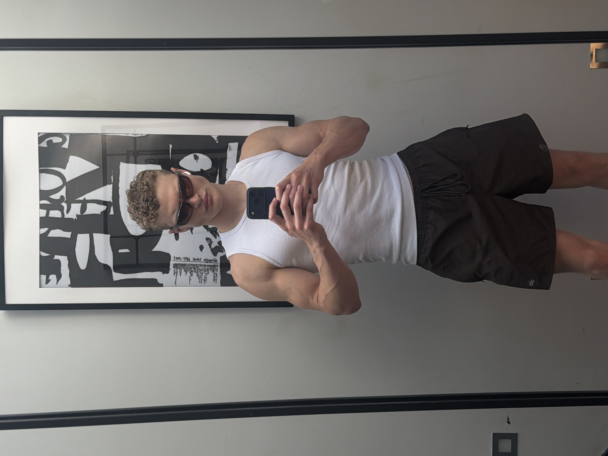 A young man with curly blonde hair and sunglasses is taking a mirror selfie in what appears to be a fitting room or dressing area, possibly in a retail store, given the presence of a framed abstract artwork on the wall and what looks like a clothing rack or partition to the left. He is wearing a white ribbed tank top and dark brown athletic shorts with a drawstring. His right arm is raised, holding a dark-colored smartphone, and his left arm is slightly bent at the elbow. The man is positioned in the center of the frame, with a neutral expression. The background is a plain, light gray wall. The overall lighting is bright and diffused. There is no indication of the specific location being within the city of London, United Kingdom.