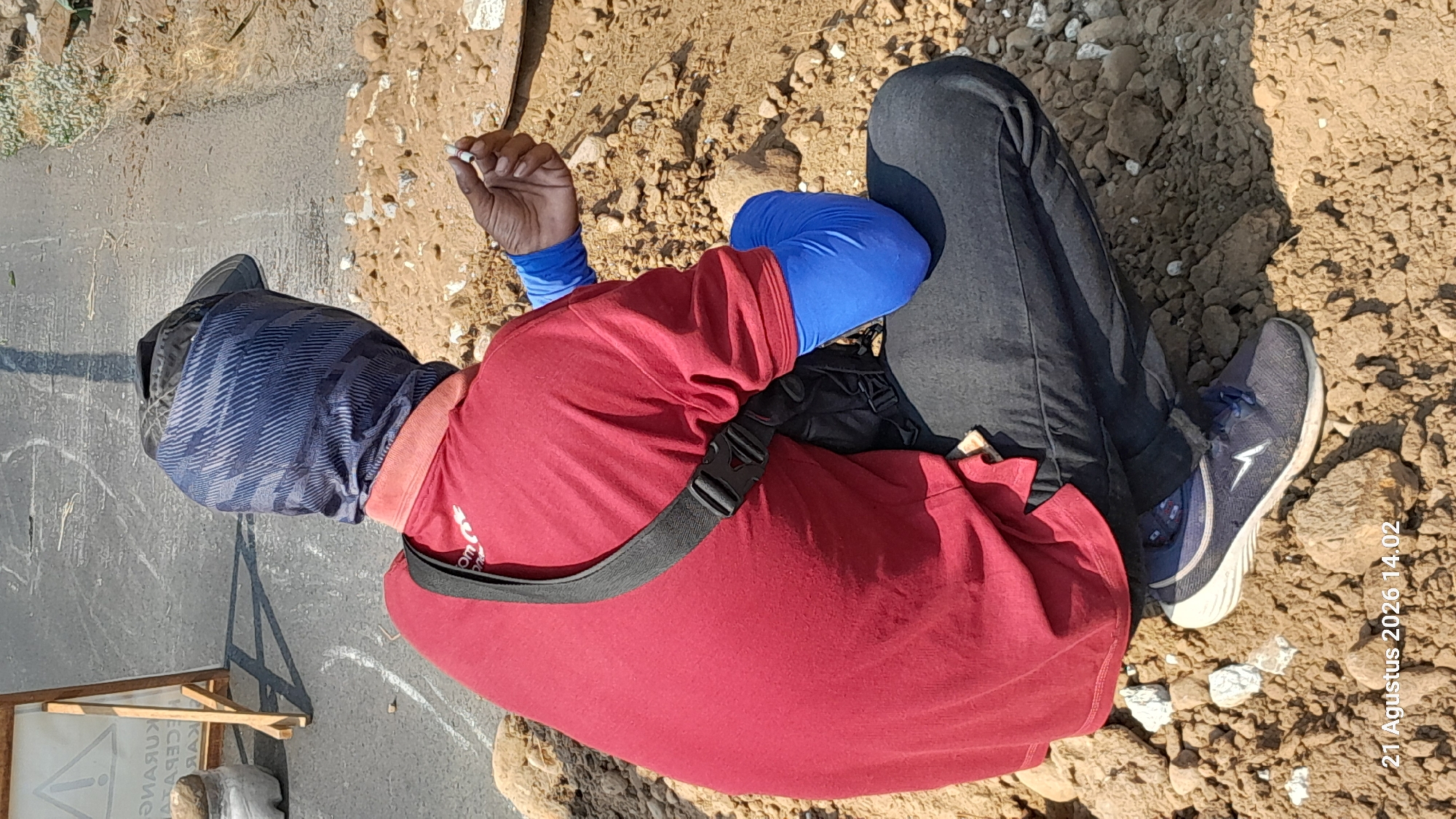 A person wearing a red shirt, black pants, a blue patterned head covering, and blue arm sleeves is crouching on a pile of dirt. The person is holding a cigarette in their right hand. A black bag is slung across their shoulder. The ground appears to be a mix of dirt and rocks. In the background, there is a paved surface that looks like a road with white markings. There is also a wooden sign with some text on it. The image has a timestamp in the bottom left corner: "21 Agustus 2026 14.02". The location is identified as Takeran, Indonesia.