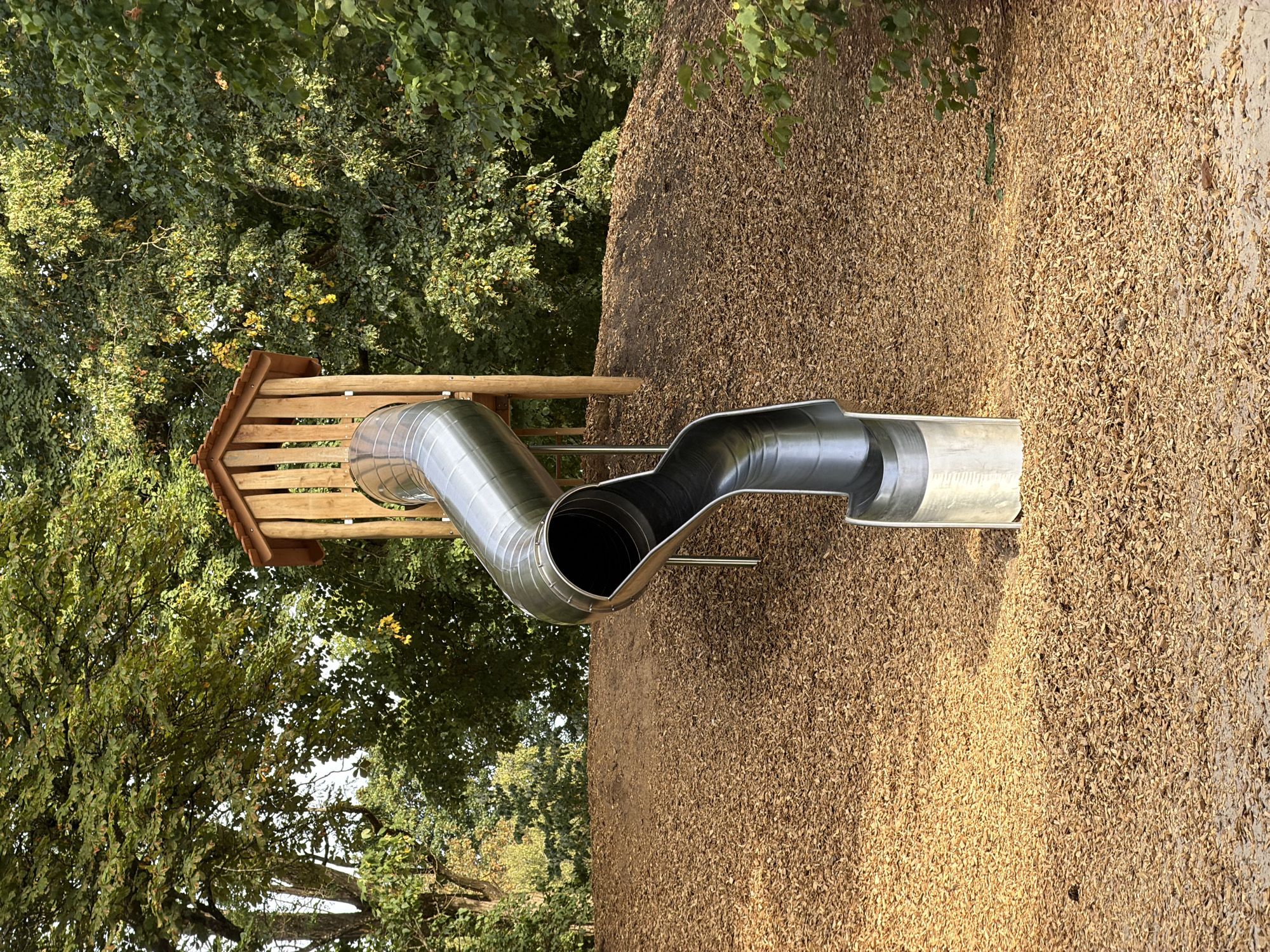 The image shows a metal slide descending from a wooden playhouse structure, which is situated on a hill covered in wood chips. The playhouse has a pitched wooden roof supported by vertical wooden posts. The slide is a curved, tubular metal structure with a wide opening at the top and a wider, flared opening at the bottom. The surrounding environment is dominated by lush green trees, suggesting an outdoor setting. The scene is devoid of any people or visible activity. This image was taken in the municipal park of Esch-sur-Alzette, Luxembourg.