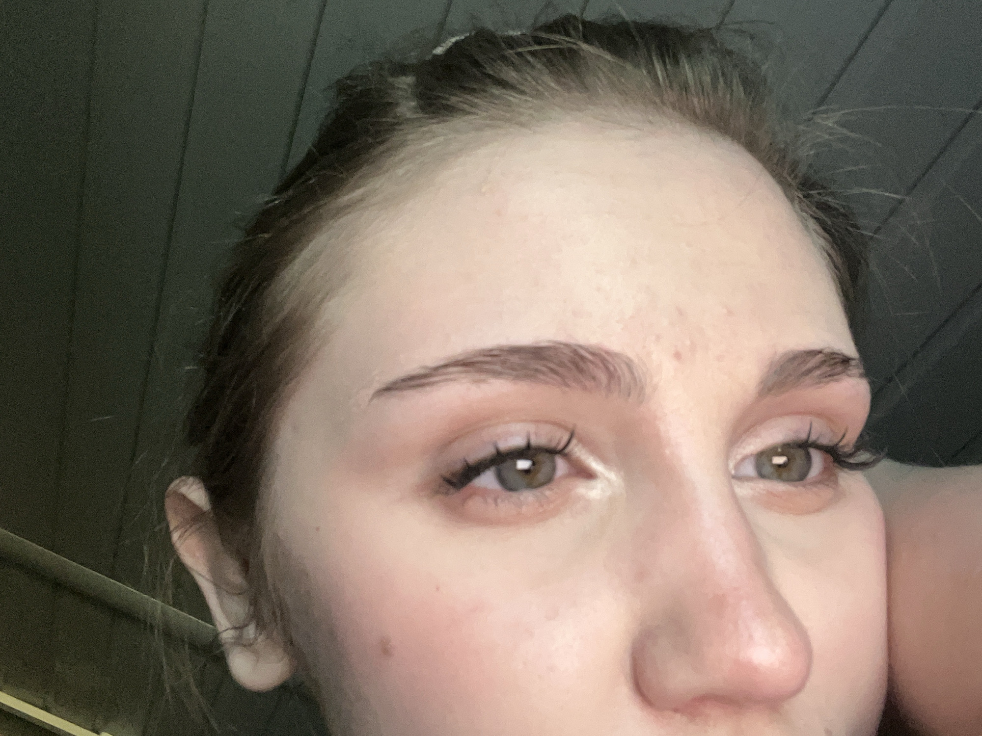 The image is a close-up, tilted, portrait-style photograph of a young woman's face, focusing on her eyes and nose. The woman has light skin with some freckles. Her eyebrows are dark and shaped, and her eyelashes appear to be enhanced with mascara. Her irises are a hazel color with visible pupils. The background is dark and out of focus, suggesting an indoor or dimly lit environment. The image is rotated, with the woman's head appearing to be facing downwards. There are no discernible city landmarks or specific geographical features visible that would indicate the location as Ashkadar, Russia.