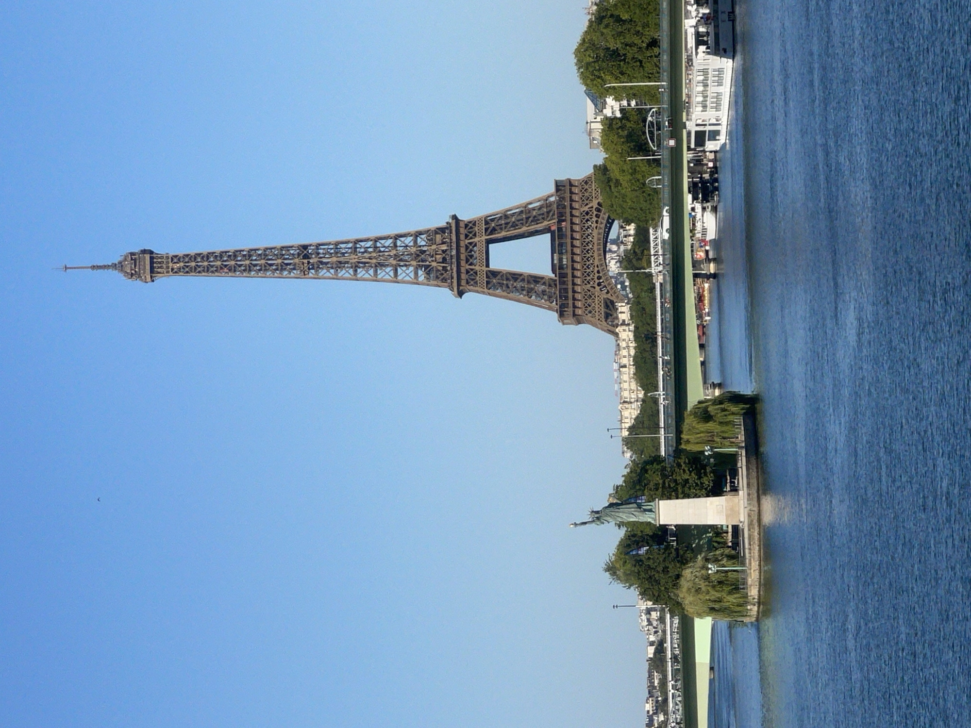 The image depicts a scenic view of the Seine River in Paris, France. In the foreground, the deep blue water of the river is visible. On the left, a small island features a statue of liberty replica, surrounded by trees and lampposts. A green bridge spans the river in the middle ground. Behind the island and the bridge, the iconic Eiffel Tower stands tall against a clear blue sky. Several buildings and lush green trees line the riverbank in the background. A few boats are docked near the bridge, and a white car is seen on the bridge itself. The overall scene is serene and picturesque, capturing a classic Parisian landscape.