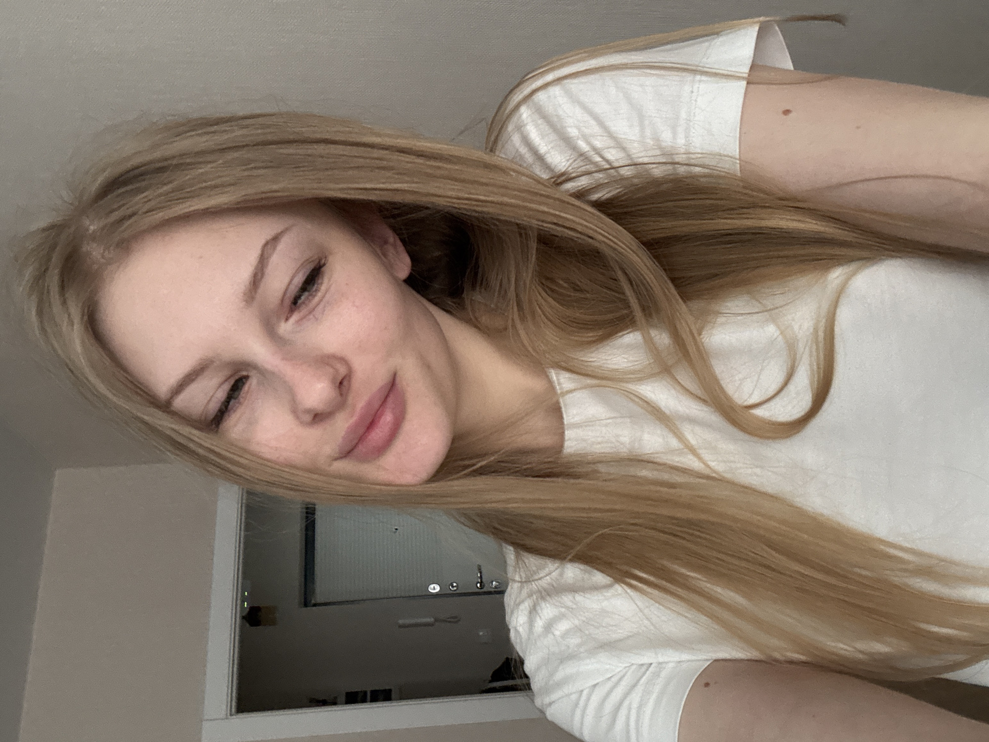 A young woman with long blonde hair is pictured in a selfie taken indoors. She is wearing a white t-shirt and her left eye is partially closed, with a slight smile on her face. She has freckles on her cheeks. In the background, a doorway and a wall are visible. The context of Perm, Russia is not evident from the visual information in the image.