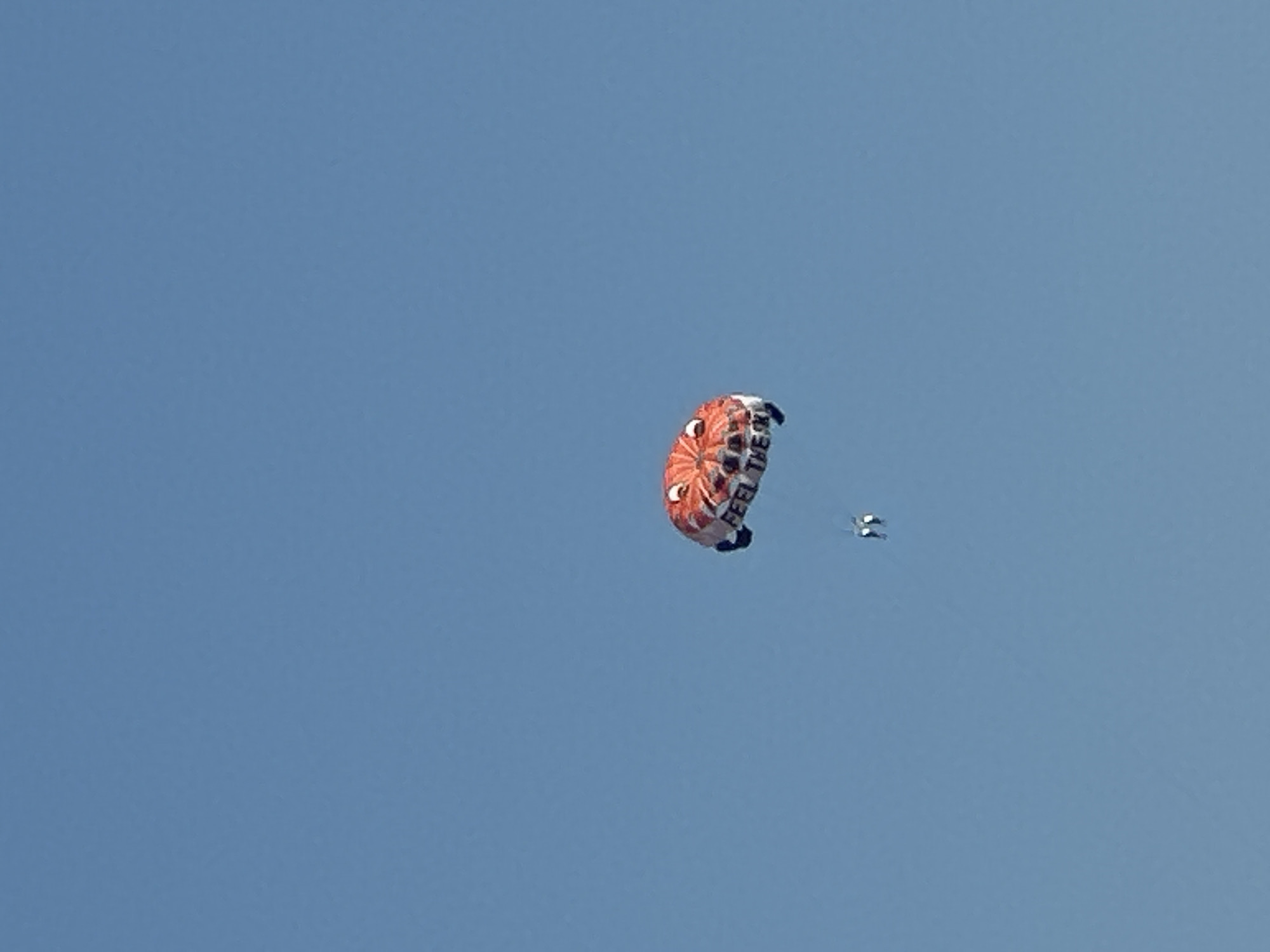 A red and white parasail designed to look like a cartoon face with large eyes and a wide mouth is in the sky. Text on the parasail reads "FEEL THE RY." Two individuals are attached to the parasail by ropes and are suspended in the air. The background is a clear blue sky. The scene is taking place in Boreti, Montenegro.