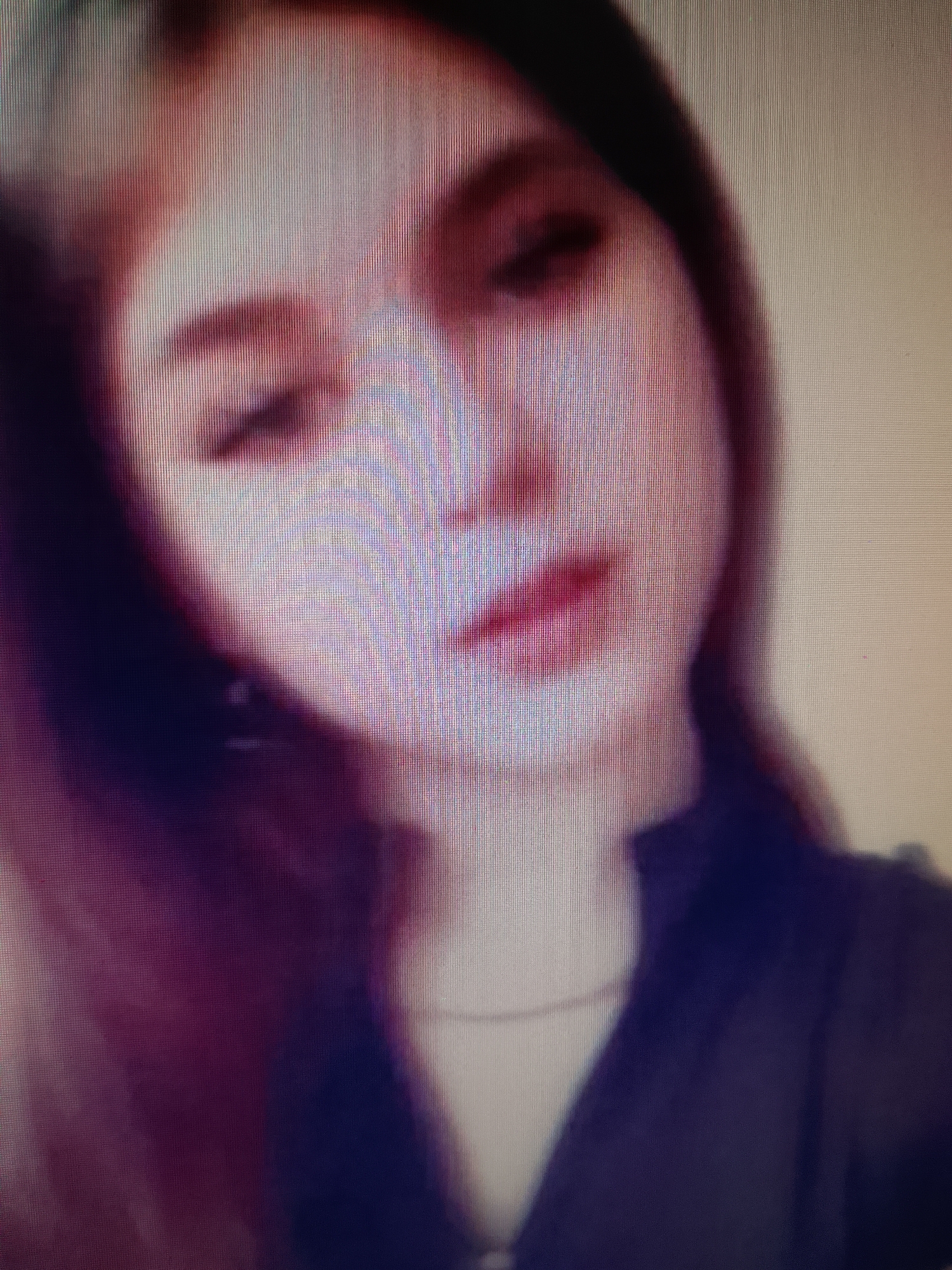 The image is a low-resolution, blurry close-up of a person's face. The subject appears to be a young woman with reddish-brown hair and fair skin. She is looking slightly to the left of the camera. Her lips are slightly parted and appear to be painted with a dark lipstick. She is wearing a dark top. The background is out of focus and appears to be a light, neutral color. There are no identifiable landmarks or features that would suggest a specific location in Ostróda, Poland.