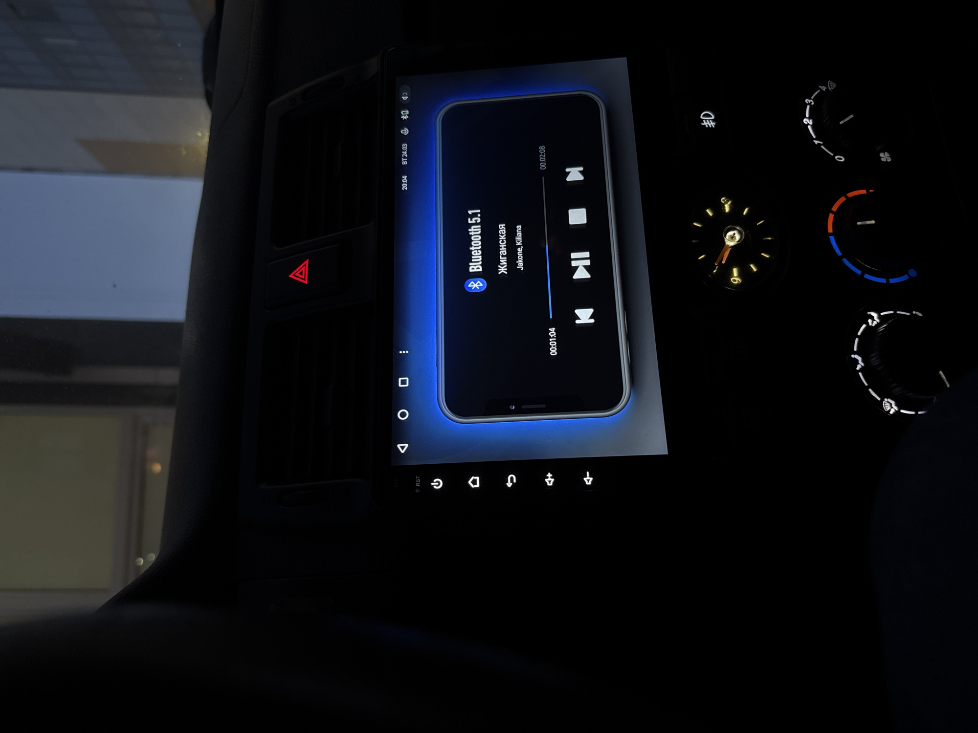 The scene depicts the interior of a vehicle at night or dusk, specifically the dashboard and a partial view through the windshield. The location is the city of Perm, Russia.
A central infotainment screen is illuminated, displaying a Bluetooth 5.1 audio connection. The song 'Жиганская' by 'Jakone, Kiliana' is playing, with a progress bar showing 00:01:04 elapsed out of 00:02:08. The screen also indicates the time as 20:04 and a date as BT 24.03. To the left of the screen, vertical touch controls for functions like power and volume are visible.
A red triangular hazard warning light button is illuminated, indicating activation. Below the screen, an analog clock shows approximately 8:00 with yellow illumination. Further below, illuminated climate control dials with blue and red segments, along with a fan speed dial, are present. A symbol for rear defogger or fog lights is also lit.
Through the windshield, the reflections of tall, modern urban buildings are visible against a dark blue sky.