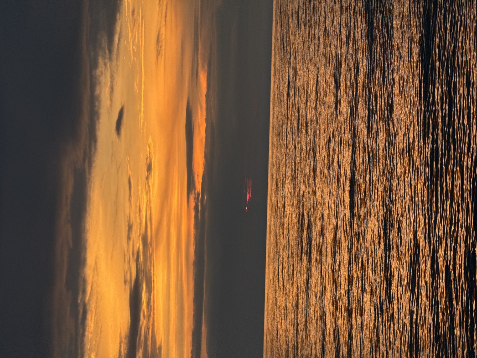 An outdoor shot of a sunset at sea, with a dark sky in the background. The sky has orange and red streaks of clouds. The sea is dark and shimmering, with a red spot in the middle. The scene is located in Batumi, Georgia.