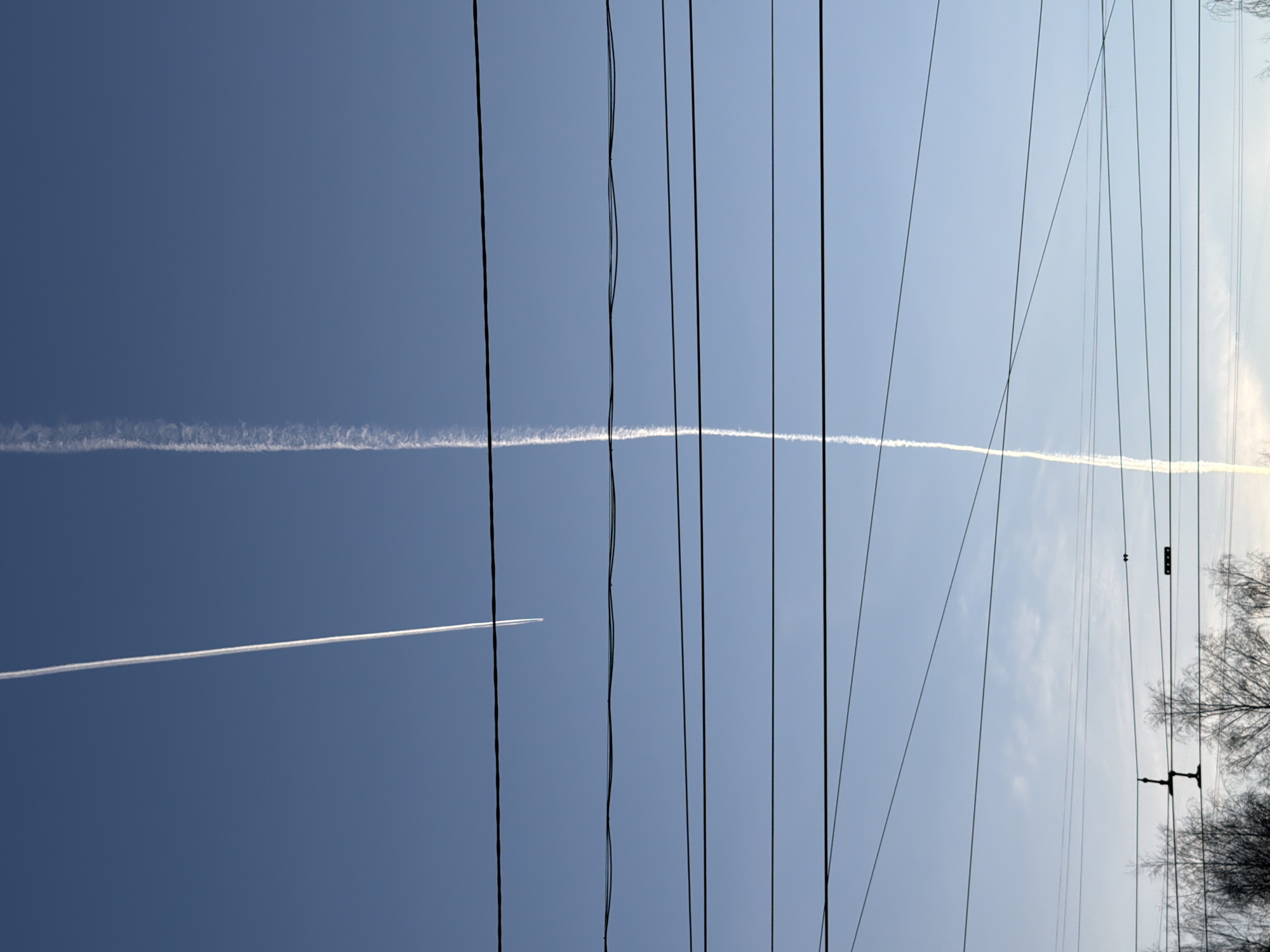 The image shows a clear, blue sky with contrails from two airplanes. Overhead power lines are present. It is unknown if the picture was taken in Izhevsk, Russia.