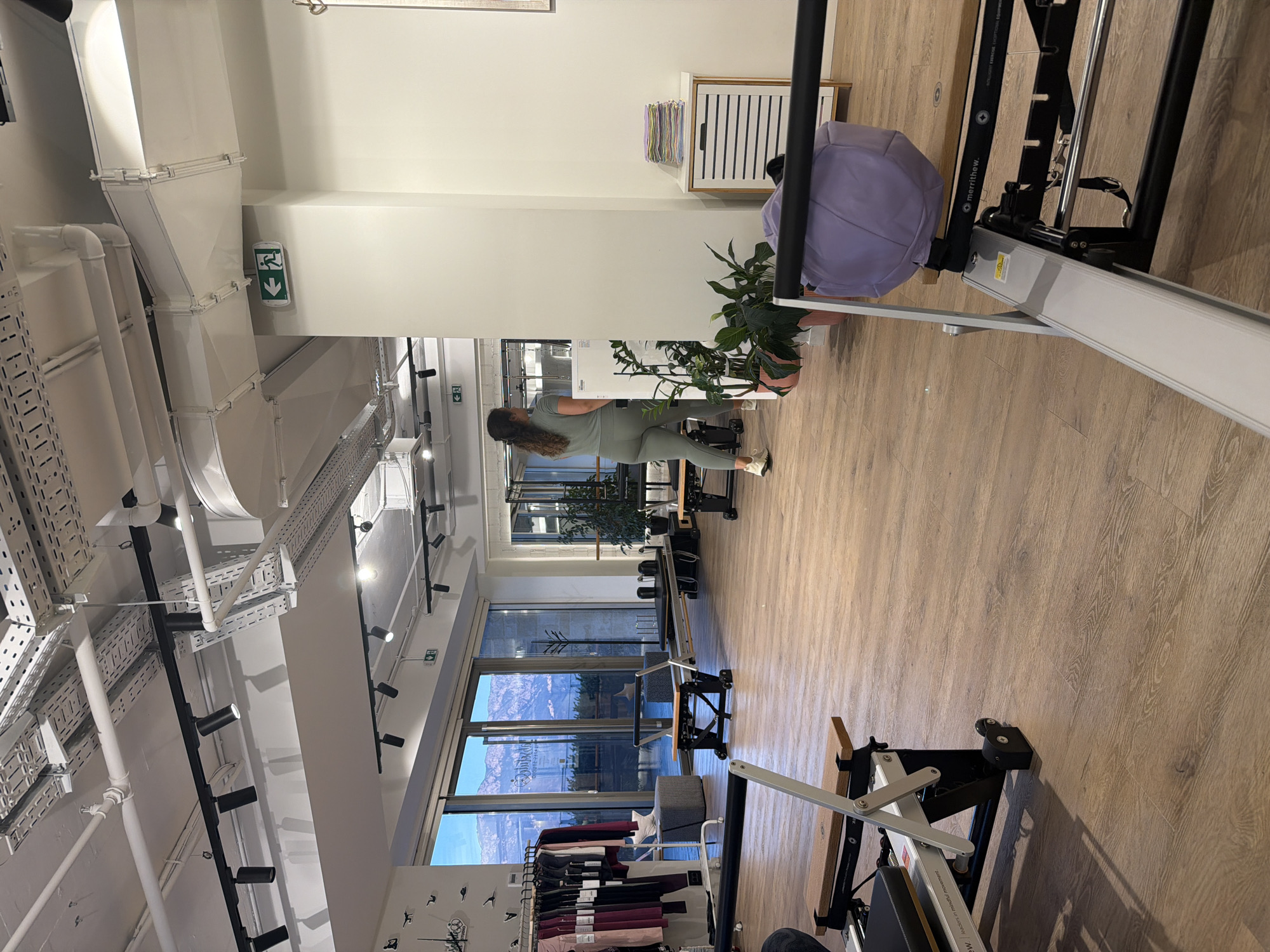 The image depicts the interior of a Pilates studio. The room has a light wood floor and a high ceiling with exposed ductwork and lighting fixtures. Several Pilates reformers are visible, some in the foreground and others further back in the studio. A woman wearing a light green athletic outfit is walking away from the camera towards the back of the studio. There is a rack of athletic clothing on the left side of the image, and large windows offering a view of mountains are also visible. On the right side of the image, there is a small cabinet with a stack of folded towels and a large purple bag. The overall impression is a clean, modern fitness space. The city of Boreti, Montenegro is not identifiable from the image.