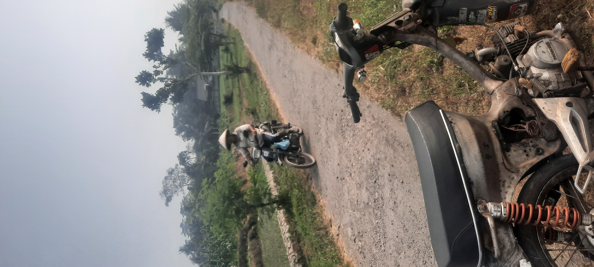 A man wearing a conical hat rides a motorcycle down a dirt road in a rural area, possibly near Ploso, Indonesia. The man is dressed in a striped shirt and dark pants. His motorcycle is dark-colored and has a blue bag attached to the front. The road is flanked by green fields and trees. In the foreground, another motorcycle is parked, partially obscuring the view of the road ahead. The sky is overcast, and the overall atmosphere is hazy. The scene appears to be a typical rural landscape, likely captured in the morning or late afternoon given the soft lighting. The presence of the conical hat suggests a Southeast Asian setting, consistent with Indonesia. The landscape indicates agricultural activity, with what appear to be rice paddies visible on the left side of the road. The condition of the parked motorcycle in the foreground suggests it might be an older model, with some visible wear and tear. The man on the moving motorcycle is focused on the road, and there is no visible interaction between him and the observer.
