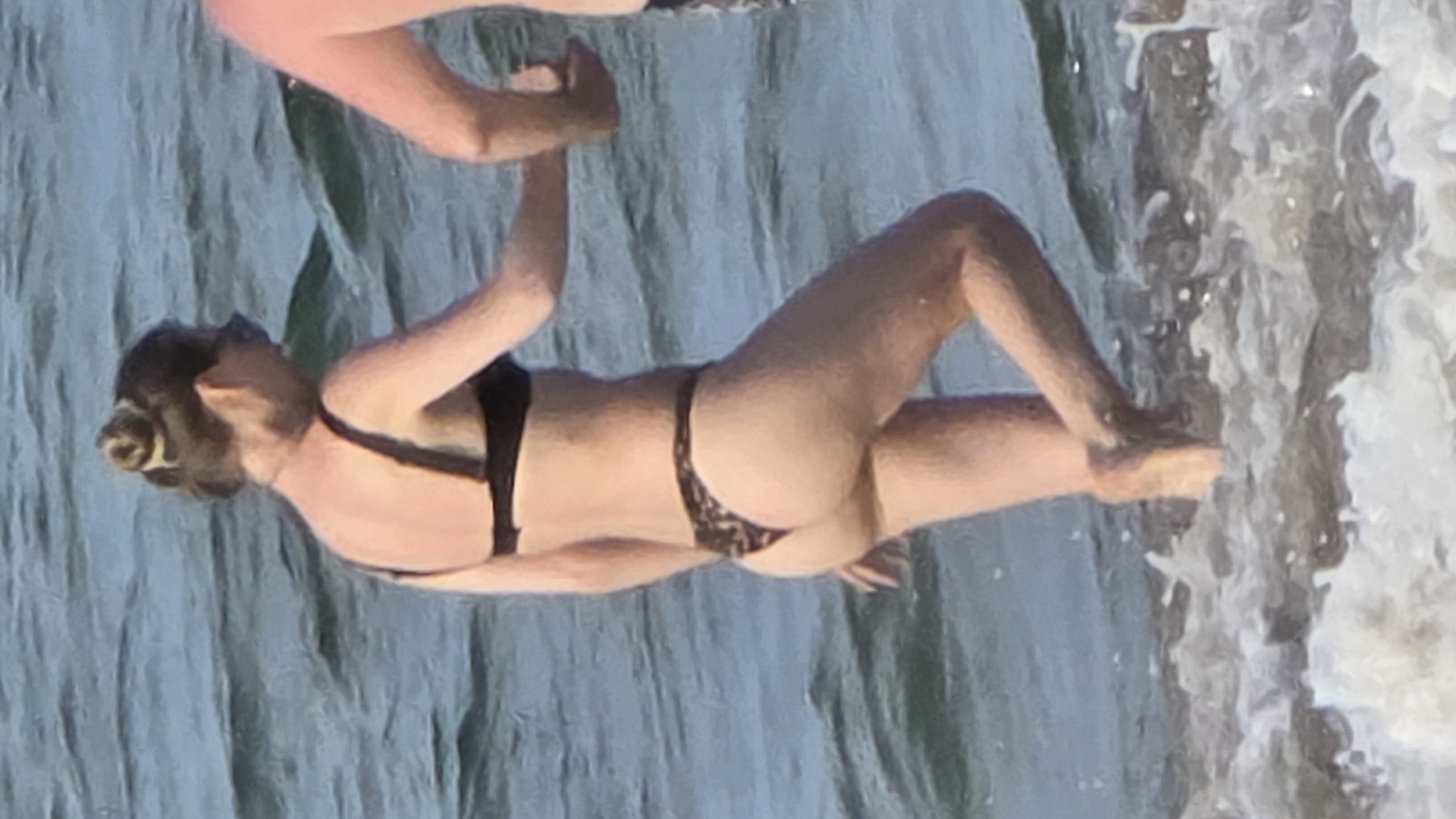 A woman in a black bikini is wading in the ocean at a beach, possibly in Nha Trang, Vietnam. She has her hair up in a bun and is holding hands with someone out of view. The ocean water is clear and light blue, with some wave action visible on the shoreline.