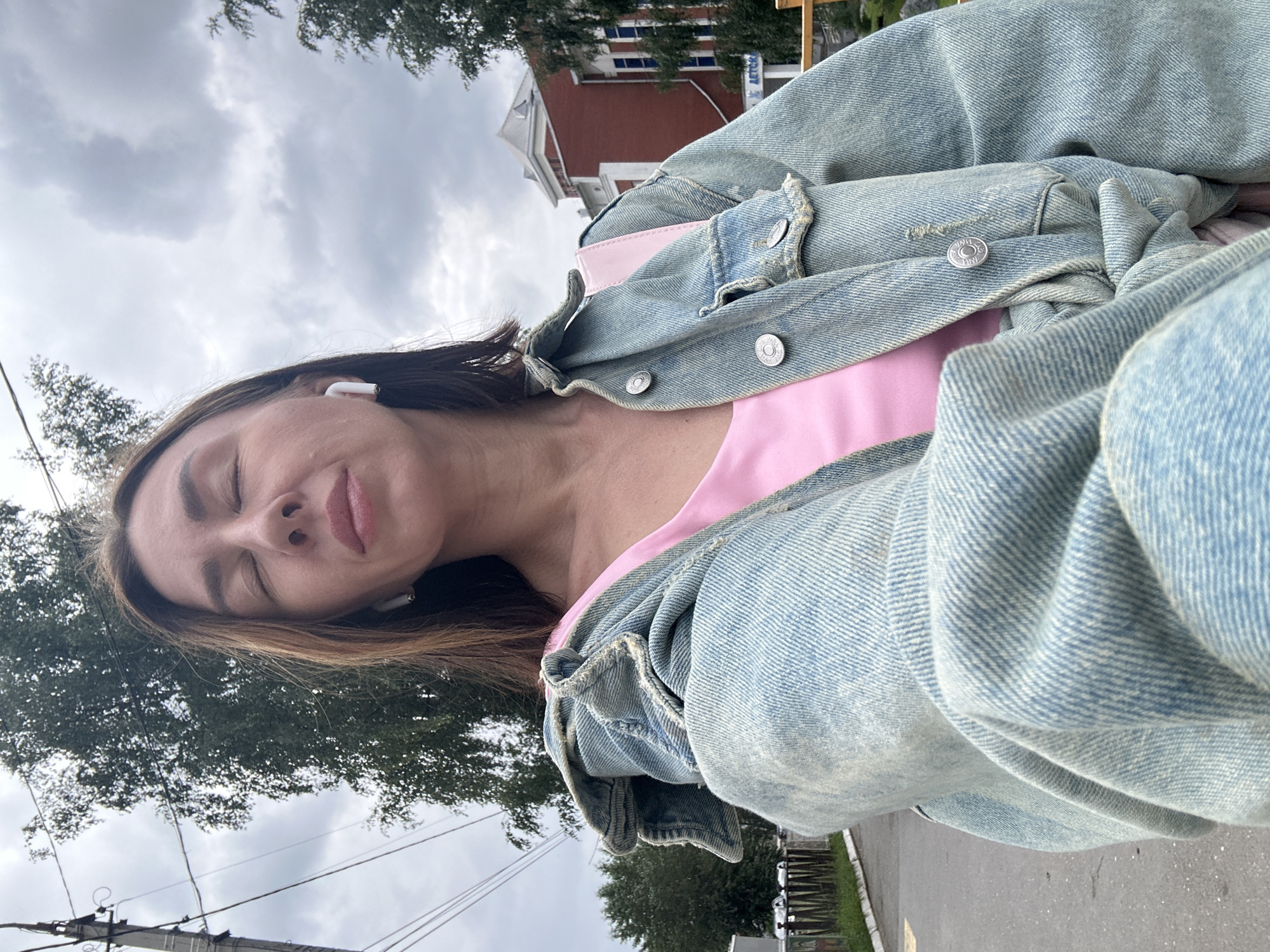A medium shot shows a woman with closed eyes, wearing a light pink top and an oversized light blue denim jacket. She has white wireless earbuds in her ears. The background is an outdoor urban scene with a cloudy sky, trees, a utility pole with wires, a building with a red brick facade and blue windows, a fence, and a paved surface that appears to be a road or sidewalk. A sign with Russian text is partially visible on the building. The overall lighting suggests an overcast day. The specific location in Votkinsk, Russia, is not identifiable from the visual elements alone.