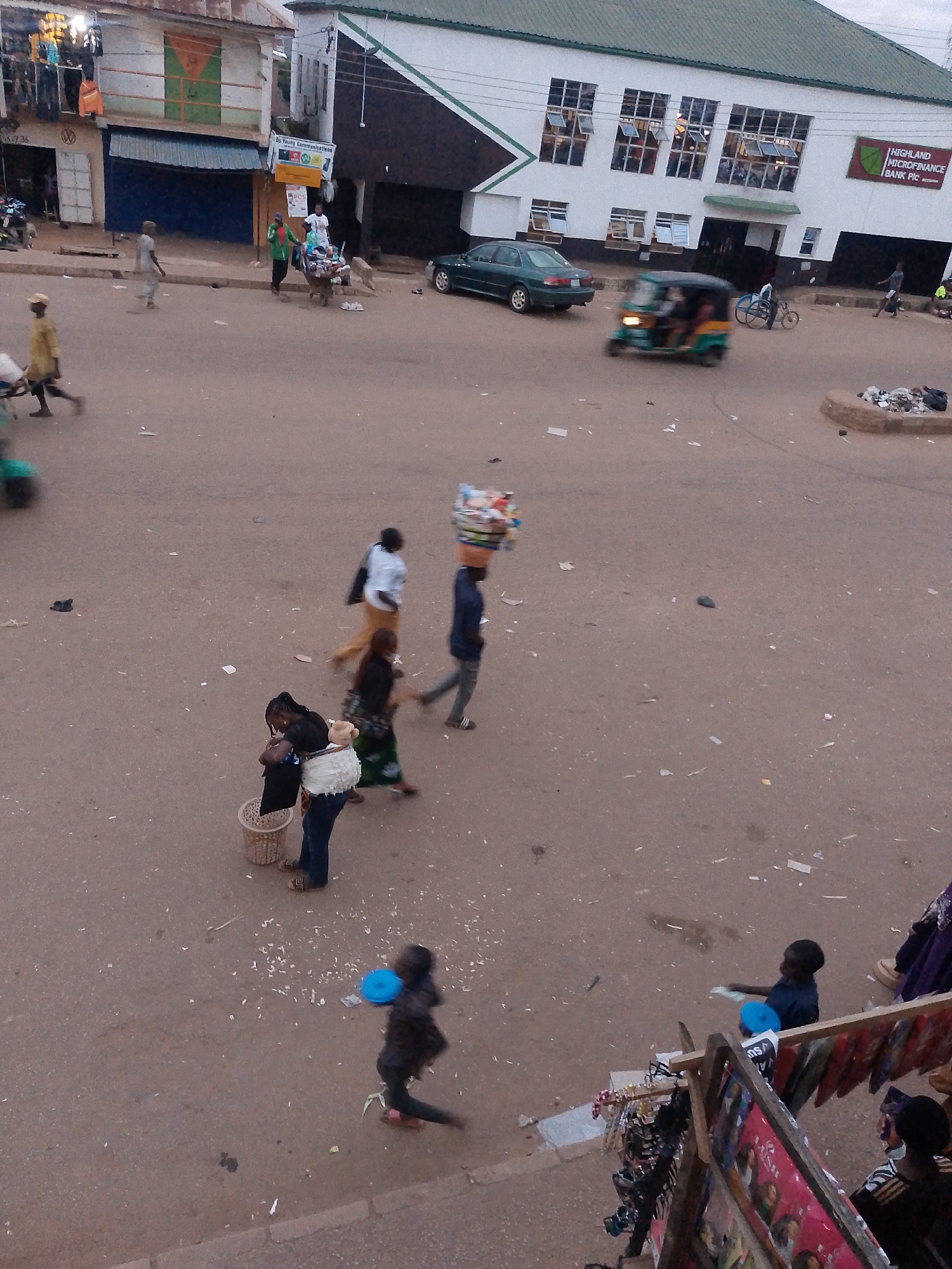 The image depicts a street scene in Bukuru, Nigeria, likely in a market area. The ground is paved with asphalt and scattered with debris. Several people are visible, engaged in various activities.
In the foreground, a child in a dark shirt is walking and holding a blue object, possibly a frisbee. To the left, a woman is carrying a basket and appears to be interacting with another person. Further up the street, several individuals are walking, some carrying items on their heads. One person is pushing a cart laden with goods.
A green and yellow vehicle, possibly a tuk-tuk or auto-rickshaw, is moving on the street. In the background, buildings line the street. One building is a multi-story structure with numerous windows. Another building has a sign that reads "HIGHLAND MICROFINANCE BANK PLC". The overall atmosphere suggests a busy, urban environment.