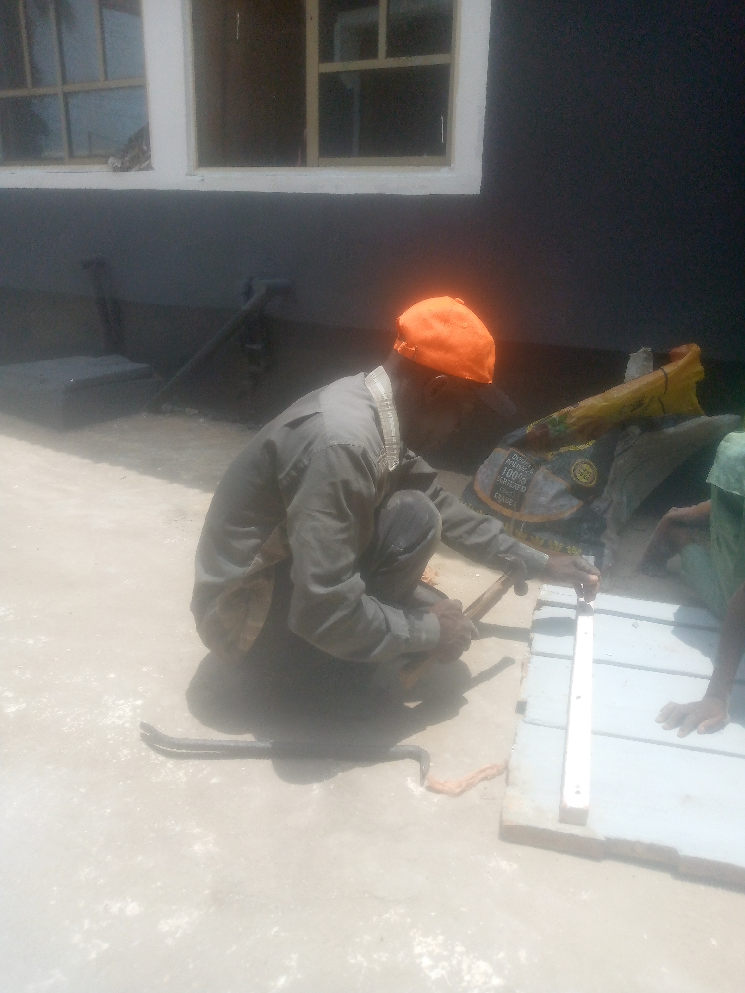 This image captures an outdoor scene featuring a man engaged in construction or renovation work under bright daylight. The primary subject is a man, possibly of African descent, squatting on a light-colored concrete or dusty ground. He wears an orange cap and a grey or khaki long-sleeved shirt, with a hint of a plaid pattern visible beneath the sleeves. He is holding a tool, resembling a hammer or chisel, and appears to be working on a grey, rectangular material, possibly a tile or block. A white straight edge or measuring stick is laid across this material.
To the man's right, part of another person's arm, clad in a light green sleeve, is visible, resting near a stack of materials. In the background, behind the working man, lies a yellow or brown sack. Text is partially visible on this sack, including "DOUBLE POLISHED," "100%," "SORTED," "GRADE," and "NET 50 KG." Further in the background, a dark grey or black building wall with white-framed windows is seen. Some pipes or conduits are also visible near the base of the wall. On the ground near the man's left, there is a crowbar-like tool.
The strong sunlight indicates it is daytime, with clear shadows cast on the ground. The setting suggests a work site or an area undergoing construction or repair, given the tools, materials, and active work being performed. The man appears focused on his task.