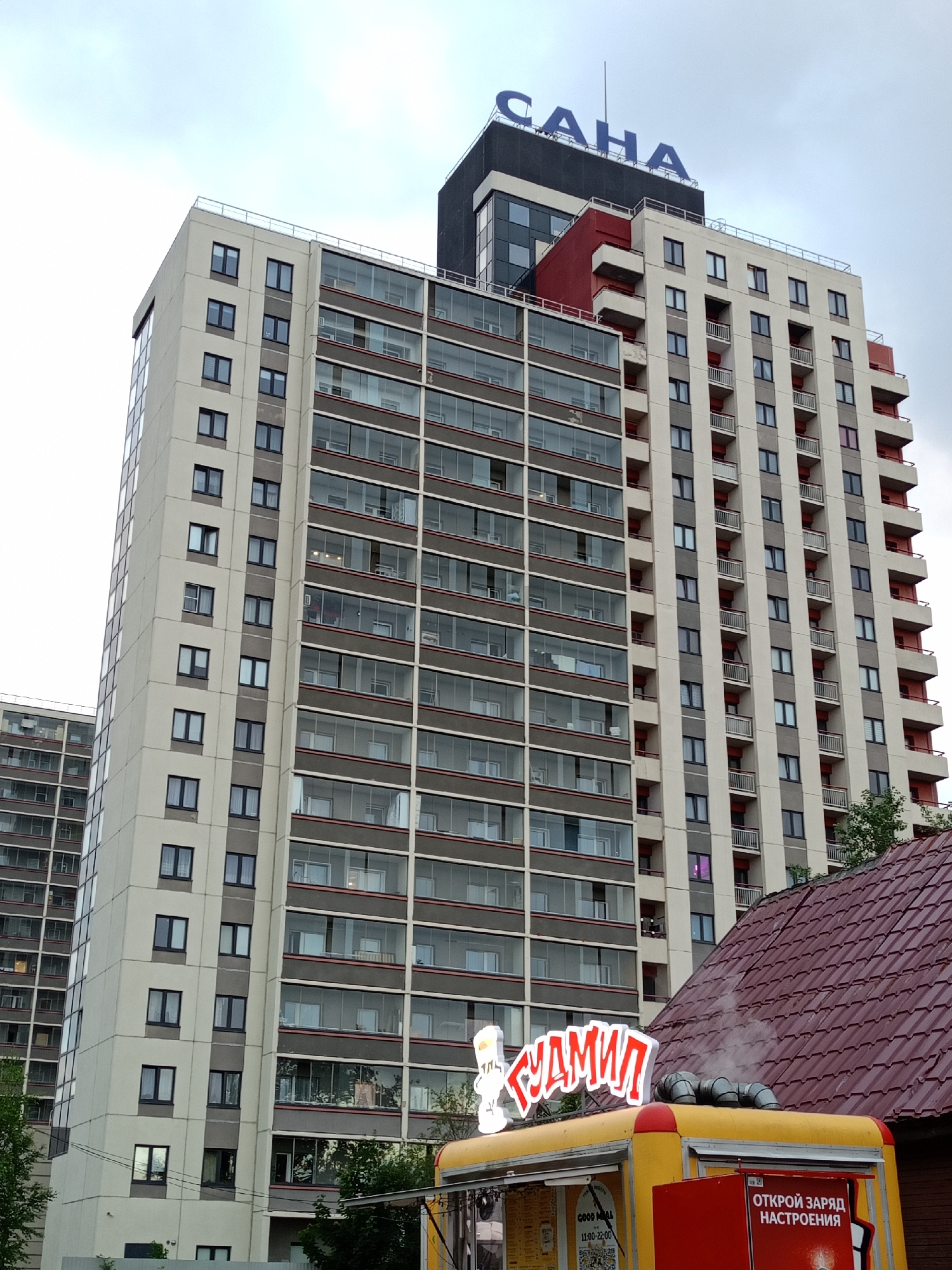 The image shows a tall apartment building with a sign on top that reads "САHA". The building is modern in design, with many windows and balconies. In the foreground, there is a yellow food truck with a red sign that says "ОТКРОЙ ЗАРЯД НАСТРОЕНИЯ". The food truck has steam coming out of a pipe on its roof. There are trees and a red-tiled roof visible in the lower right portion of the image. The sky is overcast.