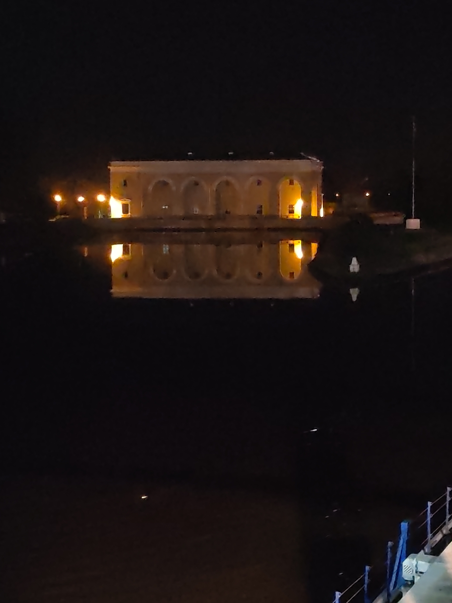 The image depicts a building with a series of arched windows or openings at night, reflected in a body of water. The reflection mirrors the building's illuminated windows and structural features. Light sources, possibly street lamps, are visible on the left side of the building. A tall pole or antenna is present on the right side. The area surrounding the water and building appears dark. According to the prompt, the location is Dedenevo, Russia. The bottom right corner of the image shows part of a blue and white fence.
