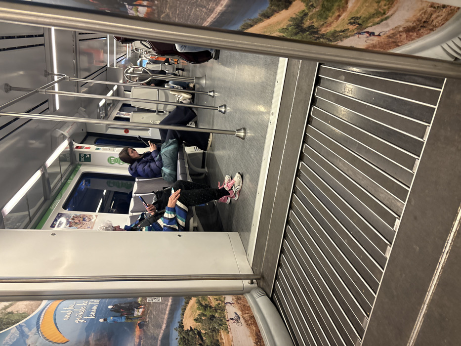 The image depicts the interior of a modern train car, likely part of the FGC (Ferrocarrils de la Generalitat de Catalunya) network, stopped at a station in Sant Quirze del Vallès, Spain. The view is from a slightly elevated perspective, looking into the main passenger compartment through an open inter-car doorway, which features a retractable metal plate with parallel slats covering the gap in the floor.
Two female passengers are prominently seated on grey, cushioned seats on the left side of the aisle. The woman in the foreground, with grey hair, wears a multi-colored striped jacket (blue, white, black), dark patterned pants, and pink and white sneakers. She is holding and looking at a smartphone. Next to her is a black bag. The woman further down the aisle has dark hair, wears a dark blue puffer jacket, dark pants, and light-colored sneakers, with her feet resting on the opposite seat. She is holding and looking at a tablet or large smartphone. A teal bag rests on the seat next to her.
Further down the train car, another passenger is partially visible on the right, seated and accompanied by a dark-colored cello in a case.
The train interior features white walls, overhead fluorescent lighting, and numerous silver vertical support poles and horizontal handrails. A red "SOS" emergency sign and other informational stickers are visible on the wall above the windows. Advertisements are displayed on the interior panels. The poster closest to the camera on the left depicts a paraglider with an orange parachute over a mountainous landscape with a lake, and a smaller image of a person cycling. Partially visible Catalan text reads "amb tu... gaudeix la tenim Tdt." A QR code is also visible on this poster. The floor inside the passenger compartment is light grey.