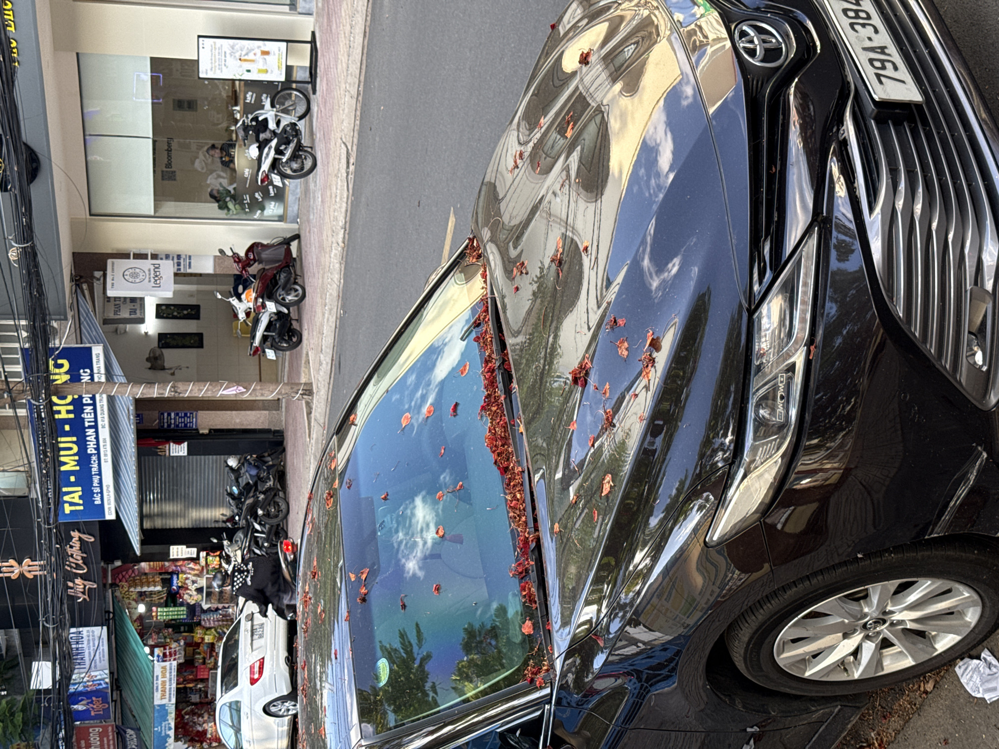 This image depicts a street scene in Nha Trang, Vietnam. In the foreground, a black Toyota Camry is parked, its windshield and hood covered in fallen red flowers. The car's license plate reads "79A-384". Behind the Camry, a white Mercedes-Benz is parked, and several motorcycles are lined up on the sidewalk. The background features storefronts with various signs, including "Thanh Hoa", "Lilly Clothing", and "Tai - Mui - Hong". A person is visible riding a motorcycle, wearing a black outfit and a patterned scarf. The environment suggests a commercial area with shops and businesses lining the street.