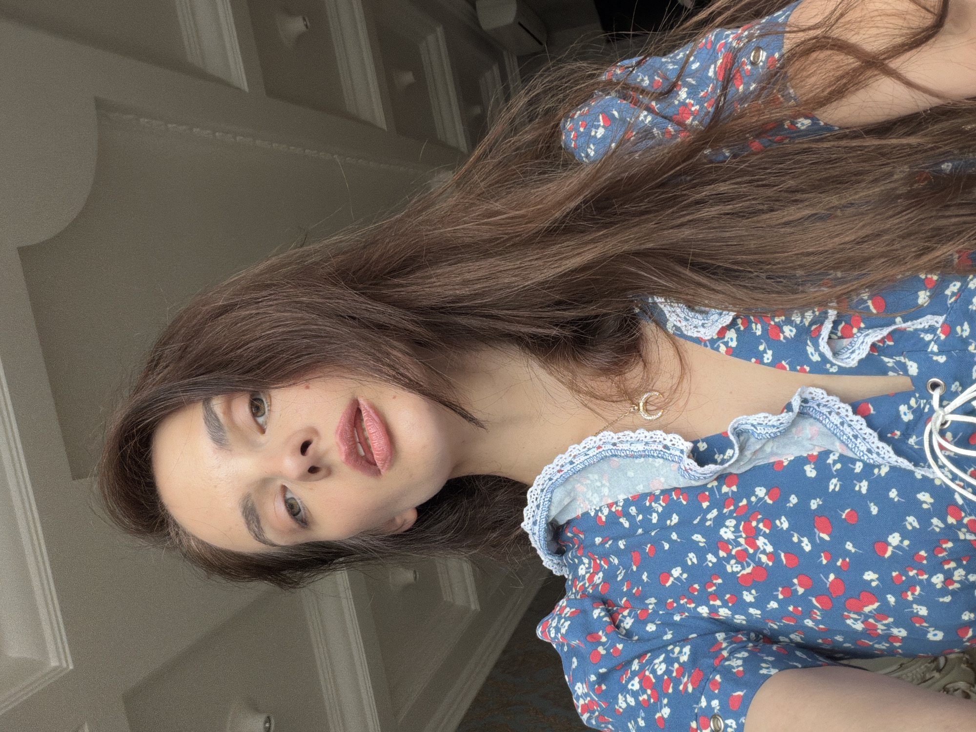 A young woman with long brown hair and light skin is taking a selfie. She is wearing a blue dress with a floral pattern of red and white flowers. The dress has a ruffled neckline and a lacing detail. She is also wearing a crescent moon necklace. The background appears to be an indoor setting with white paneled ceilings and molding, possibly in an apartment or hotel room. The lighting suggests it is daytime. There is no specific information to identify the location in Ufa, Russia.
