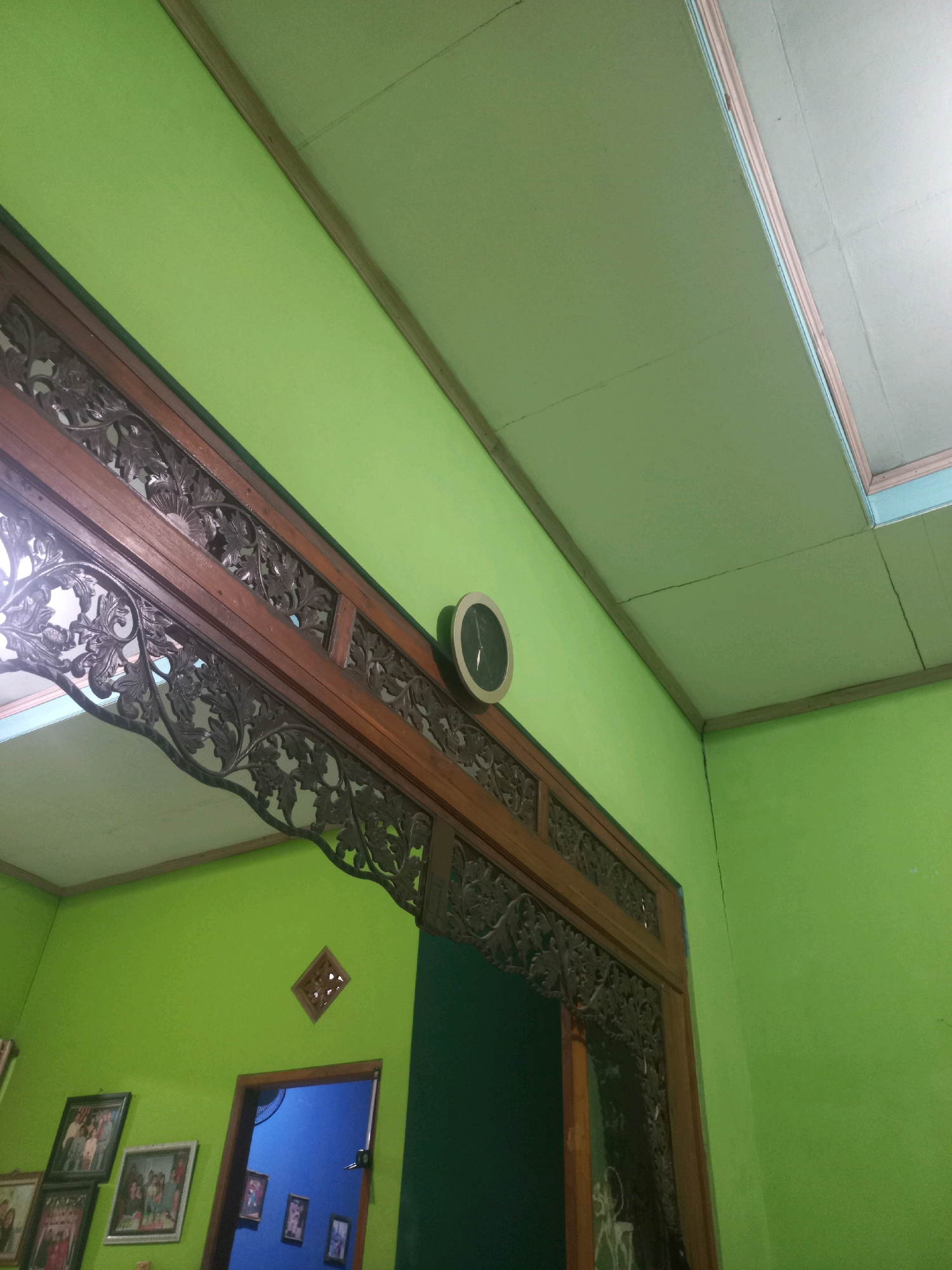 The image captures an interior view of a room with bright green walls and a light green ceiling. A dark wooden decorative archway is prominently featured, adorned with intricate carvings. To the left of the archway, several framed photographs are hung on the wall. To the right of the archway, a round clock with a black face and silver rim is mounted on the wall. A doorway with a blue interior is visible through the archway. The scene appears to be located in Sukoharjo, Indonesia.
