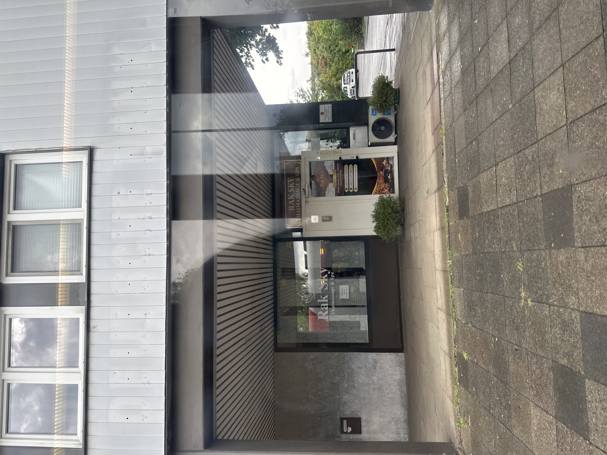The image shows the exterior of a "Rak Sky" Thai Massage & Spa establishment located in Krefeld, Germany. The building has a modern facade with horizontal paneling above the storefront. The storefront itself features large glass windows and a double door. Through the windows, signage for "Rak Sky" is visible, along with what appears to be an advertisement for Thai massage services. A white air conditioning unit is situated outside the entrance, flanked by two potted green bushes. A small, white car is parked on the street in the background, with trees and foliage beyond. The foreground consists of a paved walkway with square tiles. The overall scene suggests an urban or commercial area.