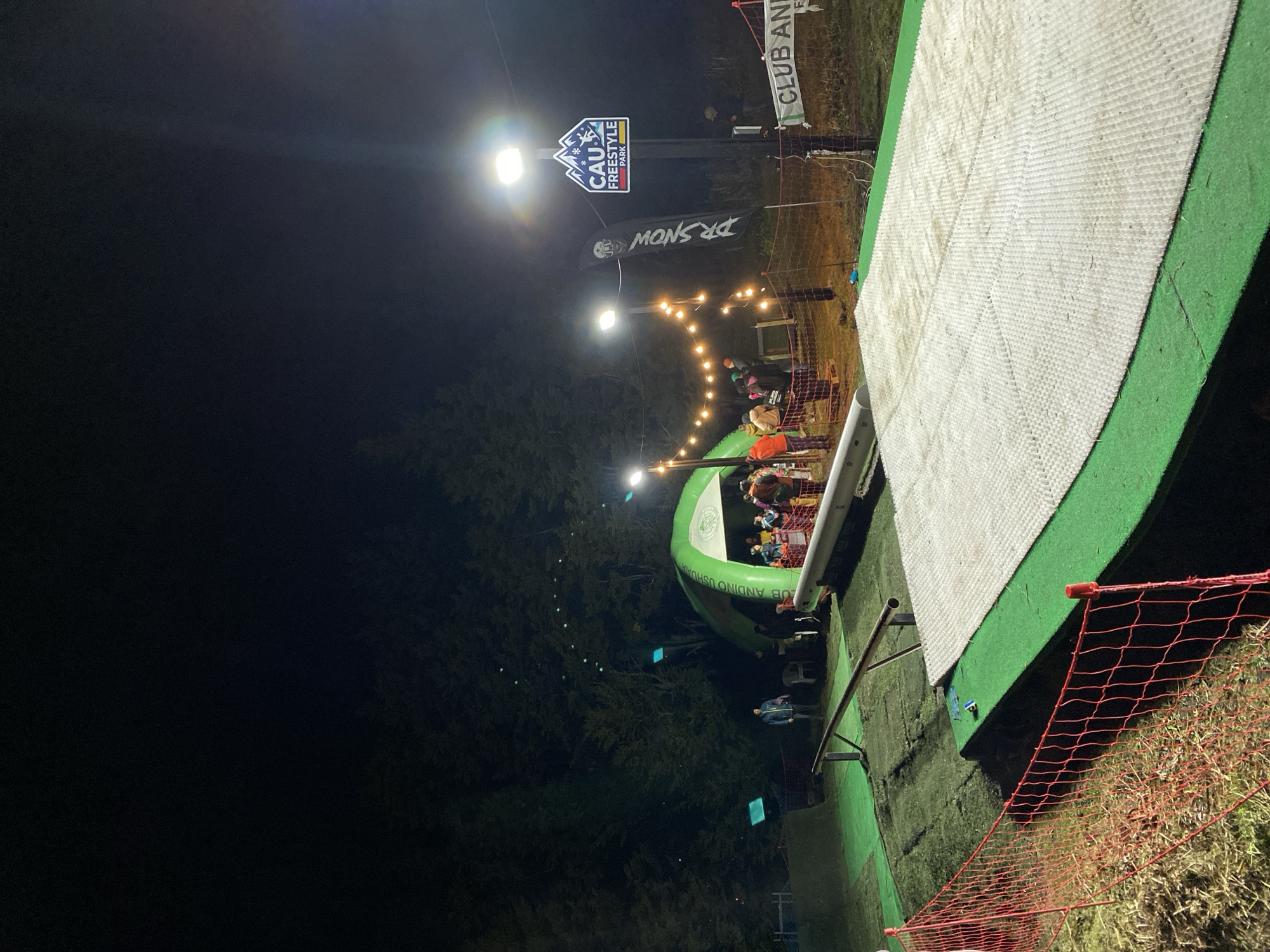 The image captures an evening scene at the CAU Freestyle Park in Ushuaia, Argentina. The focus is on a freestyle jump area with a white ramp surface and safety netting. Overhead string lights illuminate a gathering of people. Nearby, a CAU Freestyle Park sign is visible, along with a DR SNOW flag and a Club Andino Ushuaia sign. Trees flank the area, contributing to the night ambiance.