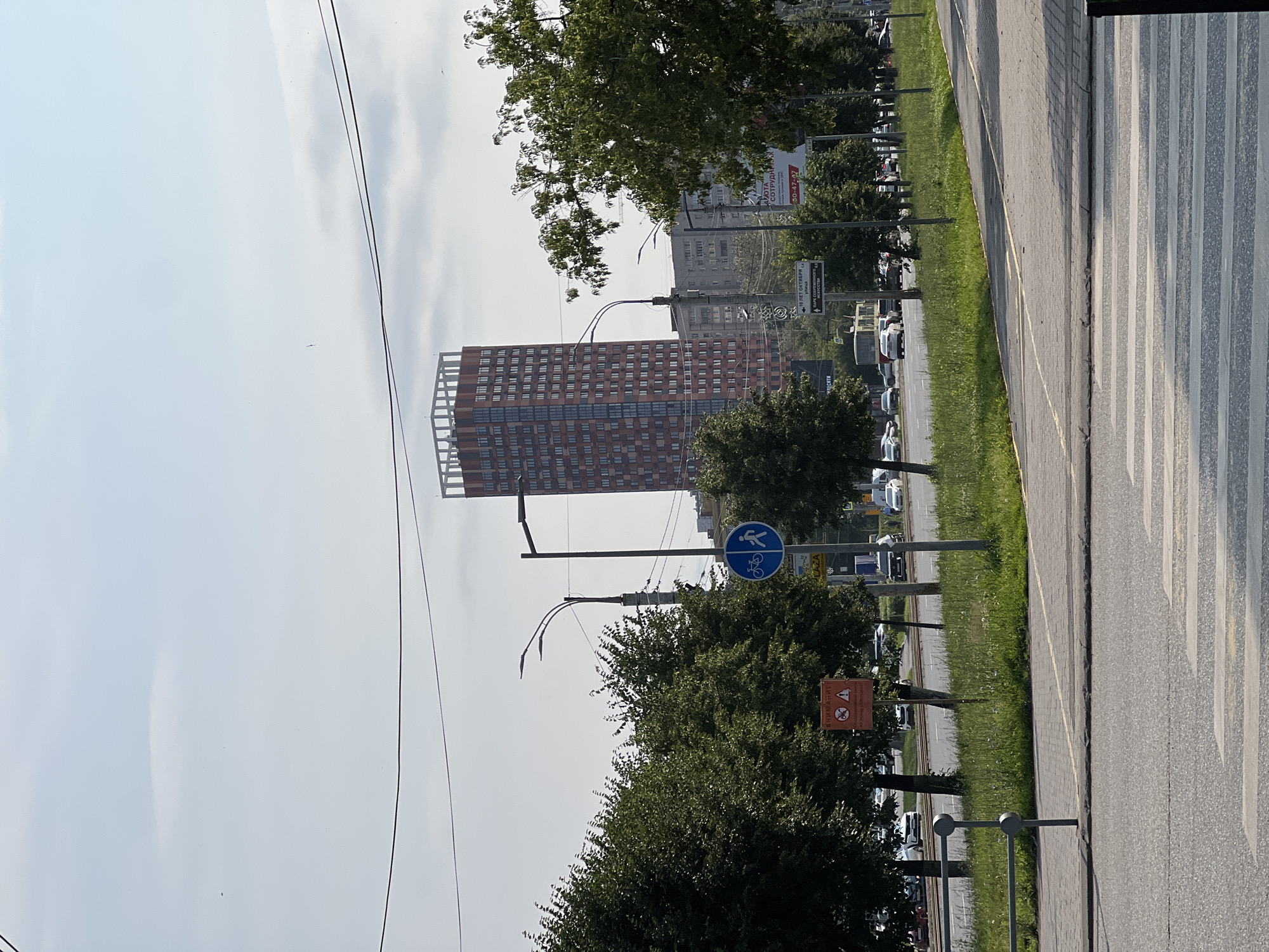 The image depicts a street scene in Izhevsk, Russia, dominated by a tall, reddish-orange skyscraper with a white lattice-like structure at its top. The skyscraper stands in the background, with other residential buildings and trees in front of it. A road with a pedestrian crossing is in the foreground, with cars visible on the road. Signs are present, including a blue circular sign with a bicycle and a pedestrian icon, and an orange triangular warning sign. Trees line the side of the road, creating a green border between the road and the buildings. The sky is light blue with some clouds. The overall impression is that of a contemporary urban environment.