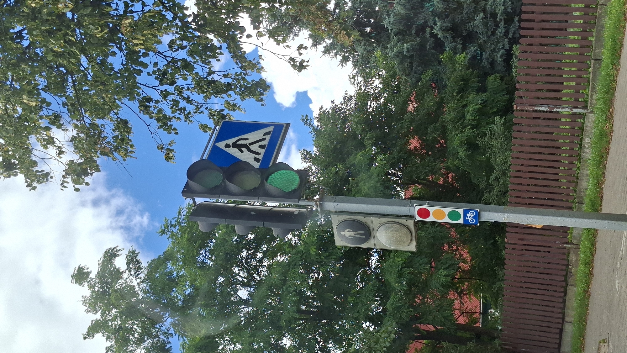 A traffic signal pole stands on a sidewalk with a brown wooden fence in the background. The traffic light system includes a standard three-light signal for vehicles, showing a green light. Below this, there is a pedestrian signal showing a walking person illuminated in white. Further down the pole, there is a secondary traffic light with red, amber, and green lights, along with a blue sign indicating a bicycle crossing. Lush green trees and a bright blue sky with scattered clouds frame the scene. The location is identified as Maladzyechna, Belarus.