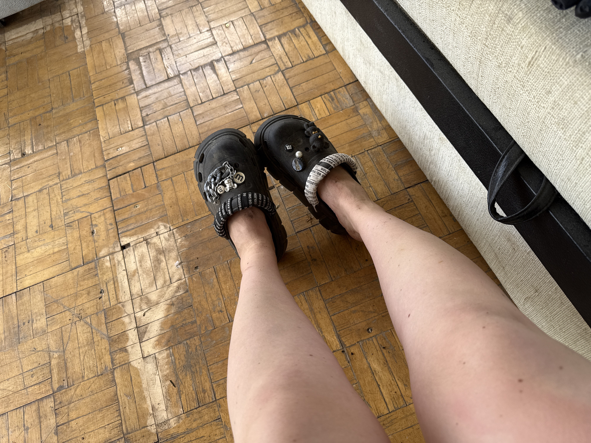 A pair of black Crocs are worn on a person's feet, with the feet positioned on a wooden floor. The Crocs are decorated with charms and fabric straps. The floor appears to be made of parquet flooring. The image does not provide any information about the location in the city of Chaikovsky, Russia.