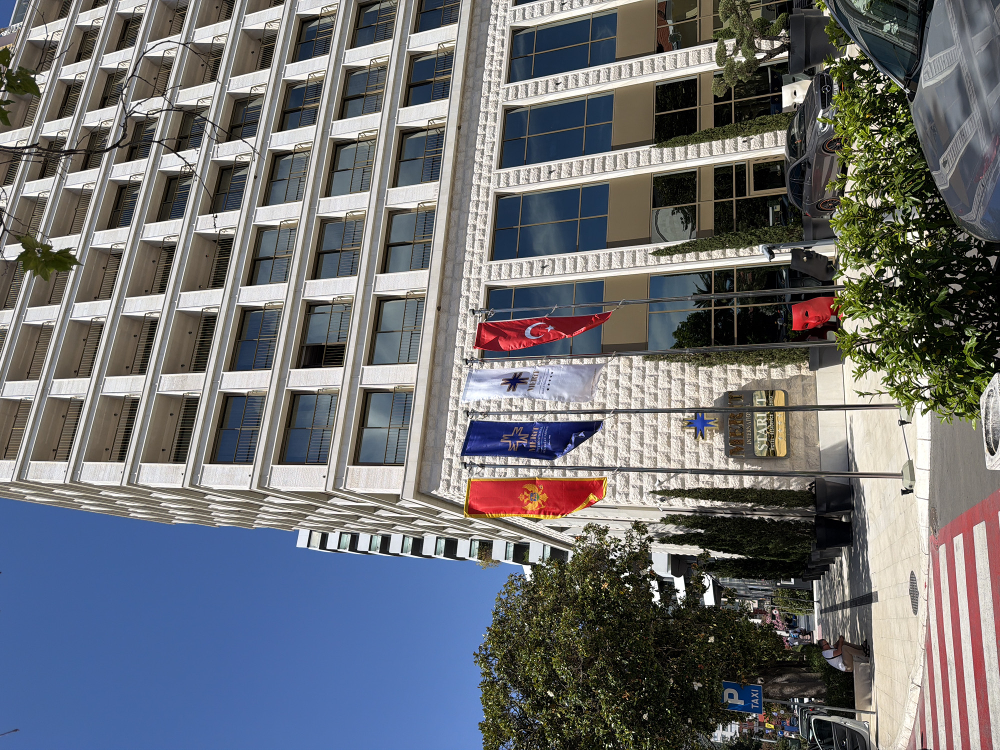 A low-angle, full-shot photograph captures the Merit International Hotel & Casino in Budva, Montenegro, under a clear blue sky. The multi-story building, clad in white textured stone, features numerous balconies with gold railings and large, dark-tinted windows. Four flags fly in front of the hotel: the red flag of Montenegro, a blue flag with a golden emblem and the text "MERIT INTERNATIONAL HOTEL & CASINO," a white flag with a similar emblem, and the red flag of Turkey with a white crescent and star.
To the left of the hotel, a lush green tree stands beside a sidewalk with a pedestrian crossing marked by red and white stripes. A blue "P TAXI" sign is visible on a pole near the tree. A man in a white t-shirt and shorts is seated on a low concrete ledge on the sidewalk, looking down. Several tall, dark green cypress trees in large black pots are arranged in a row along the building's facade.
In front of the hotel's ground-floor windows, there are several large planters containing ornamental trees. A gray Mercedes-Benz wagon is parked on the street in front of the building, and the corner of another vehicle, possibly silver, is visible in the foreground. Two artistic masks, one red and one black, are placed near the entrance. The scene depicts a sunny day in a busy urban environment with clear signs of commercial activity and international presence.