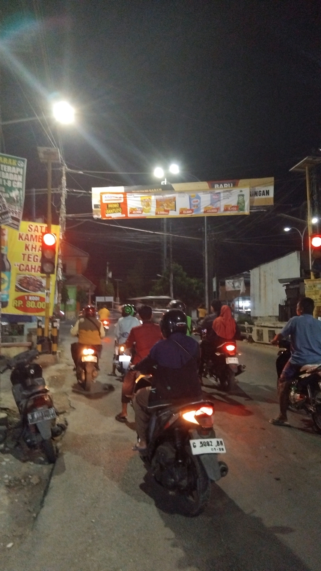 The image depicts a nighttime street scene in Tegal, Indonesia, characterized by moderate traffic and urban infrastructure. A traffic light is prominently displayed on the left side of the frame, showing a red signal, indicating that vehicles should stop. Several motorcycles are present, with their riders and passengers either waiting at the intersection or in motion. One motorcycle in the foreground has its license plate visible, reading "C 5092 BN". Billboards and signage are also visible, suggesting a commercial area. The overall ambiance is that of a busy urban intersection at night, illuminated by streetlights and the lights from surrounding buildings and vehicles.