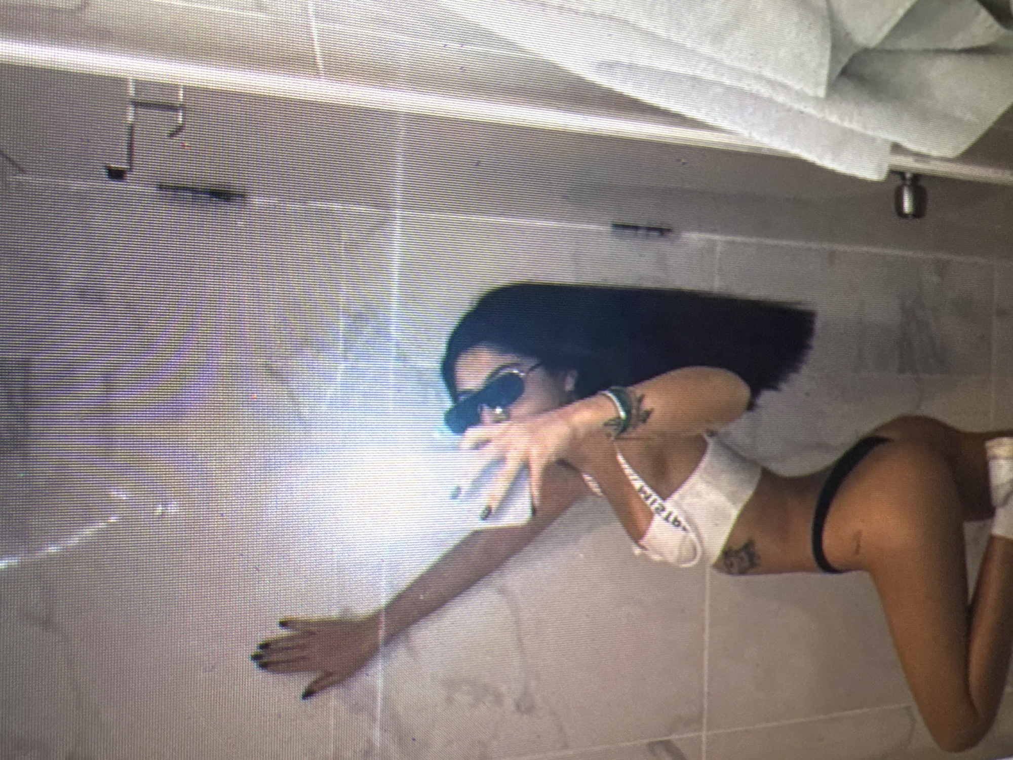 The image depicts a young woman with long dark hair and dark nail polish, wearing a white bralette and black underwear. She is posing for a mirror selfie in what appears to be a bathroom. She has tattoos on her arm and hip. In the background, there are tiled walls and a towel hanging on a rack. The lighting suggests an indoor setting. The image does not contain any information that would allow for its location to be determined, specifically in Castelo Branco, Portugal.