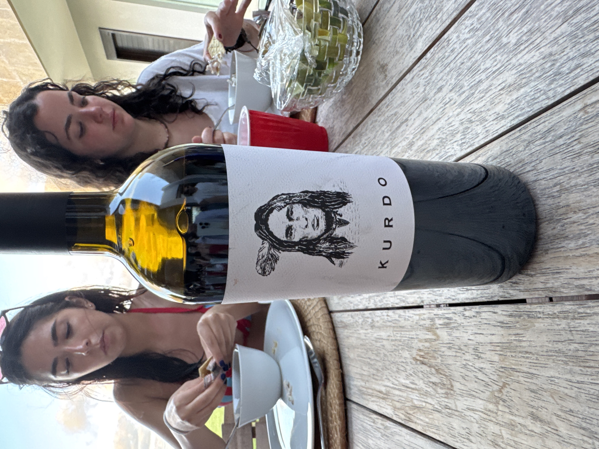 A wine bottle labeled "KURDO" is in the foreground, positioned horizontally on a weathered wooden table. The table occupies much of the image, showing visible wood grain. A young woman with dark hair, wearing a red swimsuit, is seated in the bottom left corner, looking at something in her hands. Next to her is a white plate with a cup and a spoon. Another young woman with dark, curly hair is seated in the top left, appearing relaxed. Beside her is a red cup and a crystal bowl filled with green limes. The background shows a glimpse of an outdoor scene. The location is inferred to be in or near Mata de Uva, Mexico.