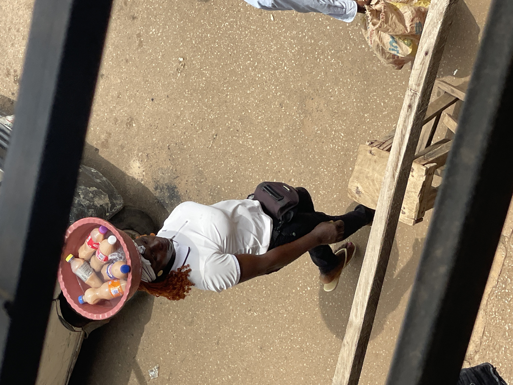 An overhead shot captures a woman walking outdoors, carrying a pink plastic basin filled with bottled drinks on her head. She is dressed in a white polo shirt, black pants, and light-colored flip-flops. A dark fanny pack is worn around her waist. Her hair is reddish and curly, and she wears a white headscarf. She is positioned in front of a dark vehicle, with a sandy, rough ground surface beneath her. To her right, a person wearing light blue clothing stands near a large, yellowish, plastic-wrapped bag. The scene appears to be in a dusty, outdoor environment, possibly a marketplace or street in Unknown, Unknown.
