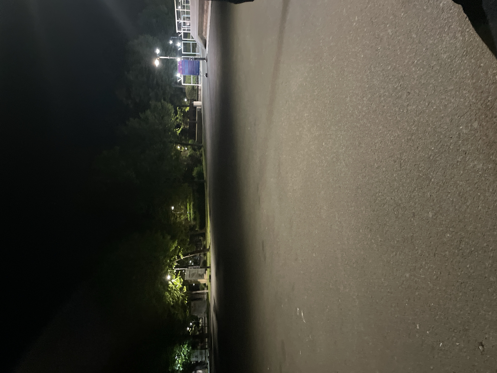 The image depicts a paved walkway in Burgas, Bulgaria at night. Streetlights illuminate the path, which is bordered by trees and foliage on one side and what appears to be a wall or low structure on the other. No people are visible in the scene. The walkway surface is asphalt or a similar material. A sign is also visible along the pathway.