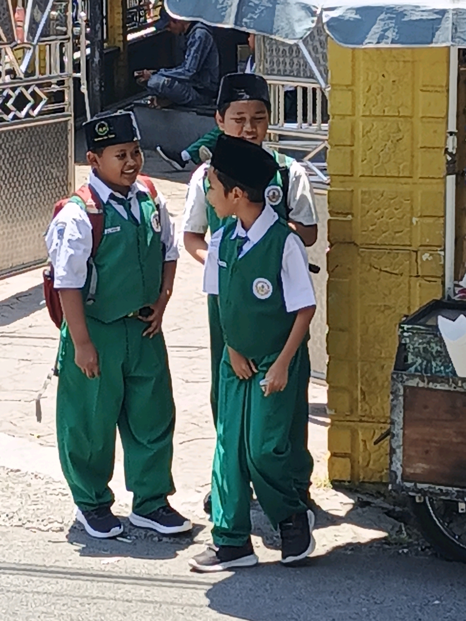 Three boys are standing on a paved area next to a yellow, textured wall. The boy on the left is wearing a black cap, a white shirt, a green vest, and green pants. He is smiling and looking to his left. A red backpack is visible on his back. The boy in the middle is wearing a black cap, a white shirt, a green vest, and green pants. He is looking forward and slightly to his right. The boy on the right is wearing a black cap, a white shirt, and green pants. He is looking forward and slightly to his left. In the background, there is a metal fence, a blue and white umbrella, and a dark wooden structure. The scene appears to be outdoors, possibly on a street or sidewalk, with sunlight casting shadows. The presence of the yellow wall and the overall setting suggest it could be a location within the city of Ketegan, Indonesia.