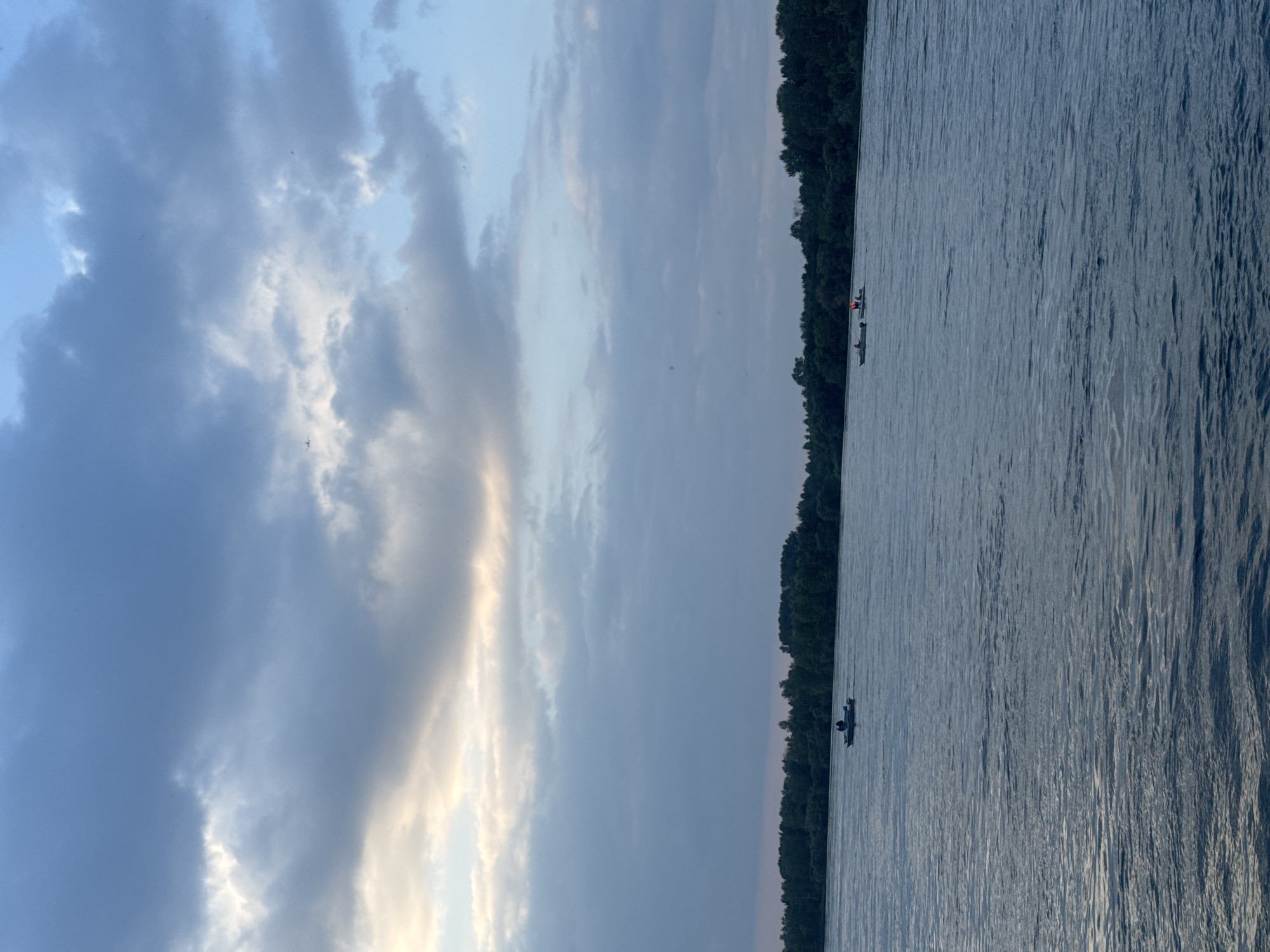 A wide, calm river dominates the foreground and mid-ground of the image. The water has a rippled texture, reflecting the overcast sky above. In the distance, a dense line of green trees forms the far bank of the river, suggesting a natural or park-like setting.
There are two small boats on the water. The boat on the left is closer to the viewer and appears to have two people in it. The boat on the right is further away and also appears to have two people, one of whom is wearing an orange life vest.
The sky is filled with dramatic clouds. Some areas are a deep blue, while others are lighter gray and tinged with a pale yellow and white, possibly indicating the presence of sunlight breaking through or an approaching sunset. A few small, dark specks in the sky might be birds or distant aircraft.
The overall impression is one of a serene, somewhat moody late afternoon or early evening scene. Given the likely location of Irkutsk, Russia, this could be the Angara River or a similarly sized waterway. The presence of boats and the natural landscape suggest an outdoor recreational area or a section of the river used for transport or fishing.