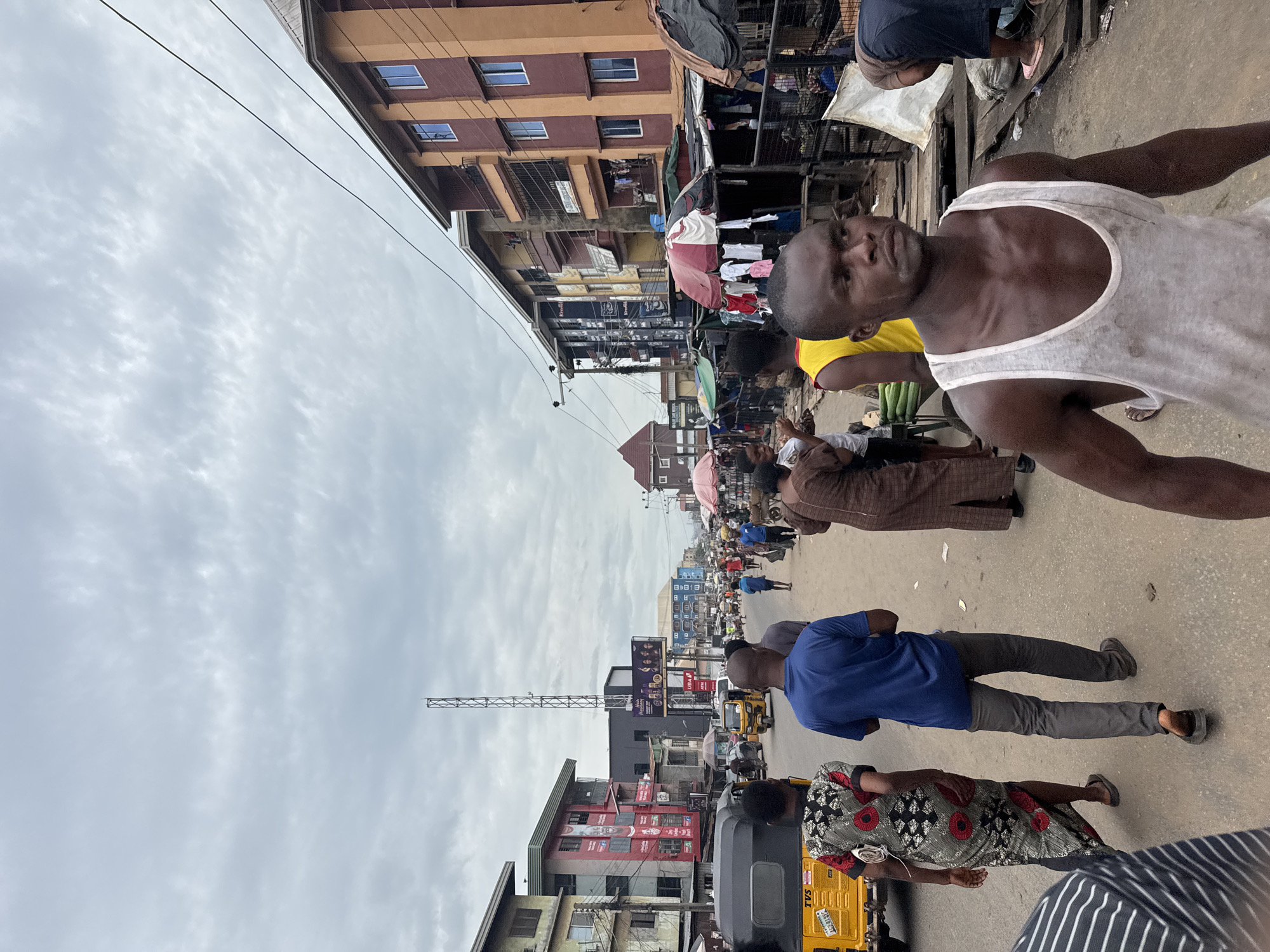 The image depicts a crowded street scene in Fegge, Nigeria. A man in a white tank top is prominent in the foreground. Many people are present, including a person in a blue shirt and someone in a patterned dress. Transportation includes a yellow auto-rickshaw. Buildings line the street, with banners and signs visible. Overhead wires crisscross the sky, which is overcast. A tall communications tower is in the background.