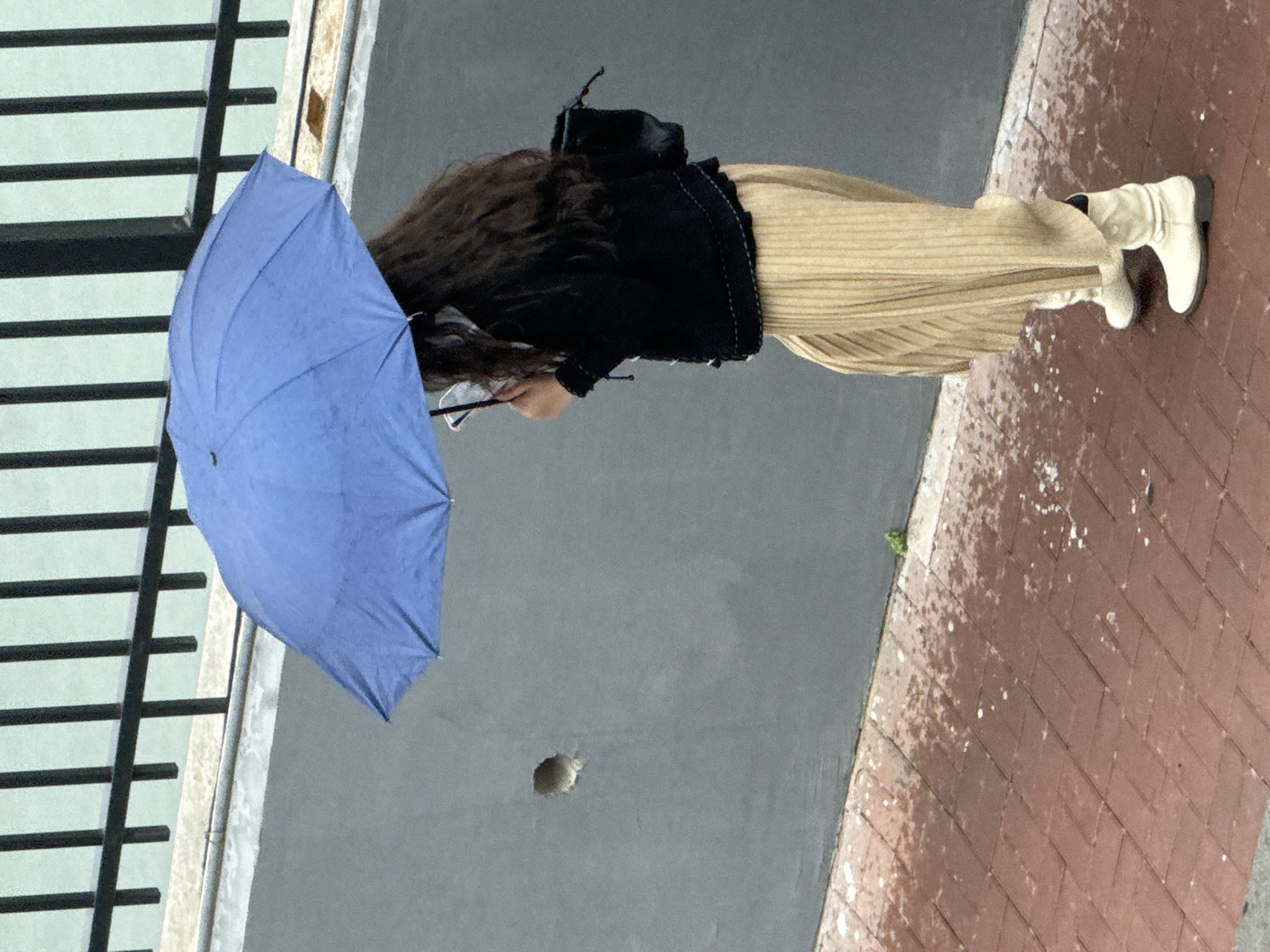 A person stands on a wet, brick-paved sidewalk next to a grey wall in Zhongtun, China. They hold a blue umbrella, wear a black jacket, a tan corduroy skirt, and white boots. A black backpack is visible. A black metal fence is to the left. A hole is visible in the grey wall.