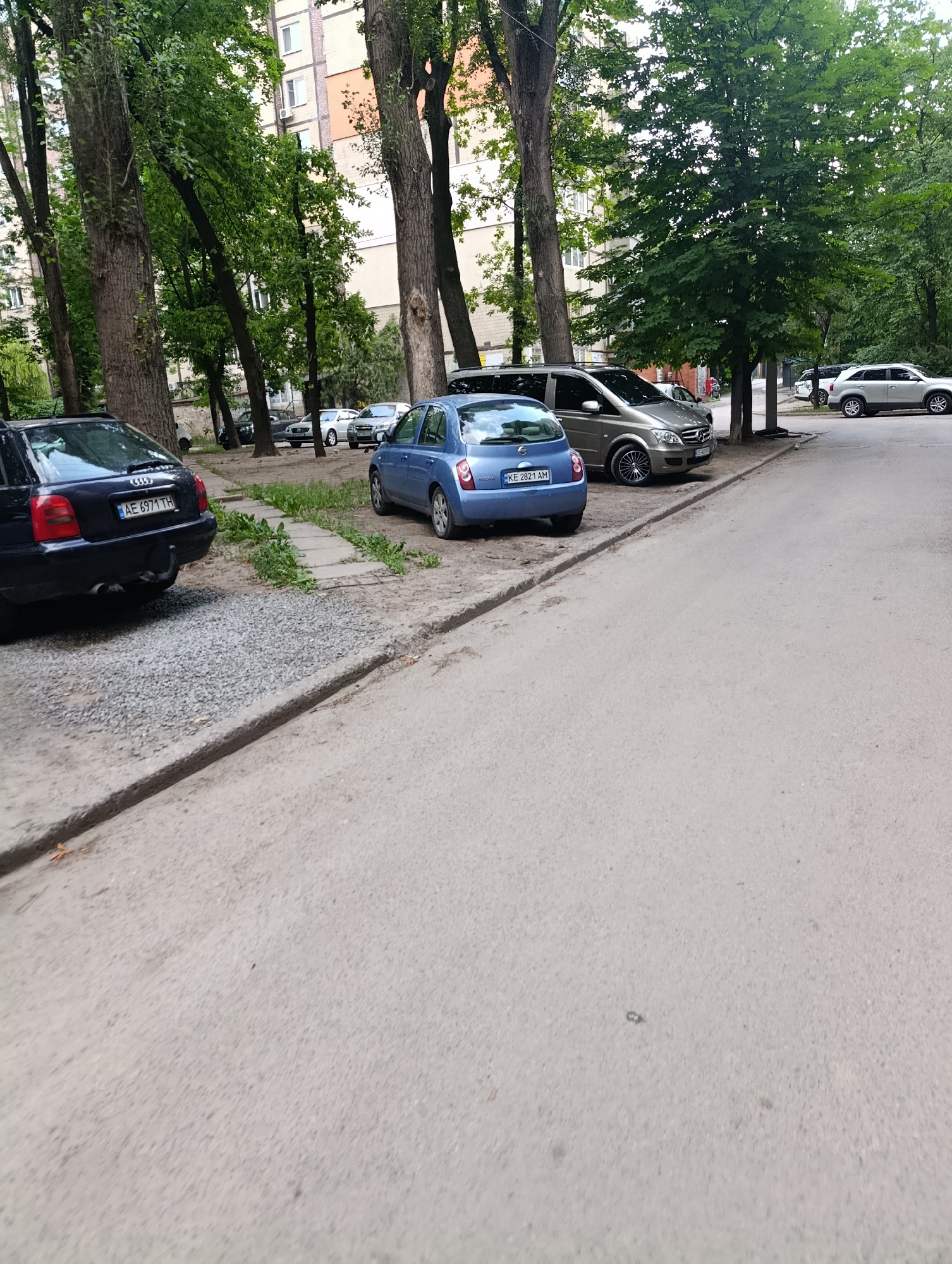 In Dnipro, Ukraine, a residential street is depicted with parked cars on the side of the road and a building in the background. The scene includes several vehicles: a dark blue Audi A4 Avant with the license plate AE 6971 TH, a blue Nissan Micra with the license plate KE 2821 AM, and a silver Mercedes-Benz Vito van. Further down the street, a silver SUV is visible. The environment is characterized by mature trees lining the sidewalk and a paved road. The ground is a mix of asphalt and gravel. The building in the background appears to be a multi-story apartment complex with cream and orange colored walls. The sky is overcast.