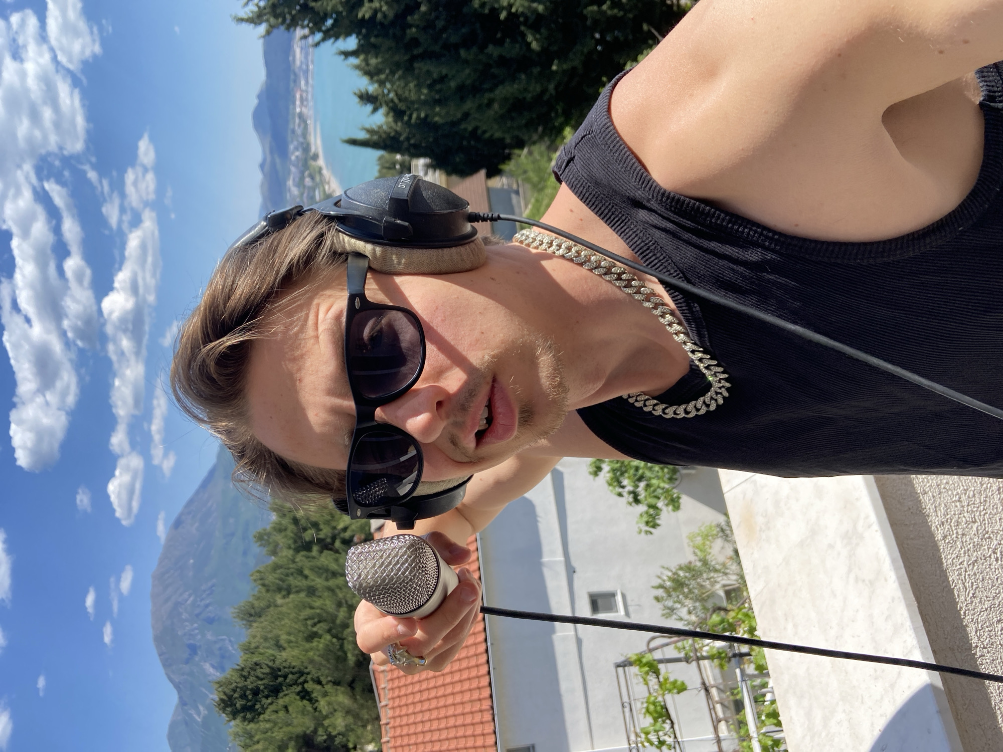 A young man is prominently featured in a sunlit outdoor scene, captured in what appears to be a selfie or a direct address to the camera. He is seen from the chest up, leaning into the frame, and looking directly towards the viewer. He wears a black ribbed tank top, dark sunglasses, and large over-ear headphones, clearly marked "DT 770 PRO" on the left earcup, with tan ear pads. A thick, silver-colored Cuban link chain necklace adorns his neck. In his right hand, he holds a silver and white wired microphone, and several rings are visible on his fingers.
The background reveals a stunning coastal landscape, indicative of Šušanj, Montenegro. Under a bright blue sky dotted with scattered white clouds, a large body of turquoise water, likely the Adriatic Sea, stretches to the left. A mountainous coastline with green trees and scattered buildings is visible in the distance. The immediate lower background shows white and orange-tiled rooftops, suggesting the man is on a balcony or rooftop overlooking the town. The scene is brightly lit by strong daylight, indicating a clear and sunny day. The man's expression is direct and focused, suggesting he is performing or recording.