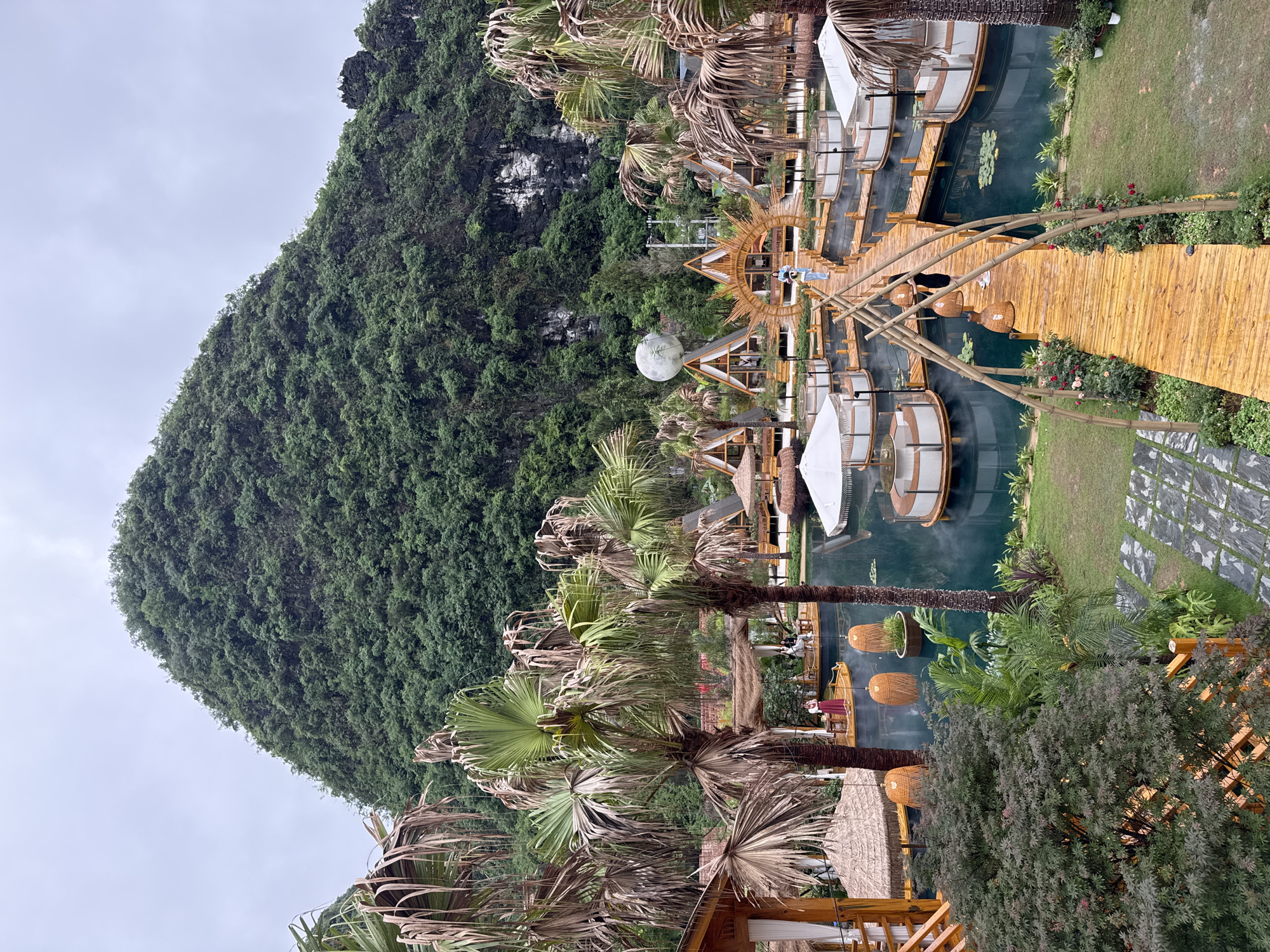 This outdoor shot captures a unique lakeside resort in Guilin, China. A verdant, tree-covered mountain dominates the left side of the frame under an overcast sky. A series of interconnected wooden platforms and walkways extend across the water. Circular, pod-like structures with white canopies are built atop these platforms. People are visible inside the structures, although obscured, suggesting that they are seated. Several decorative, lantern-like objects hang above the wooden pathways. A walkway extends into a garden area with stone paths on the right side of the image. Palm trees and other tropical foliage surround the lakeside structures.