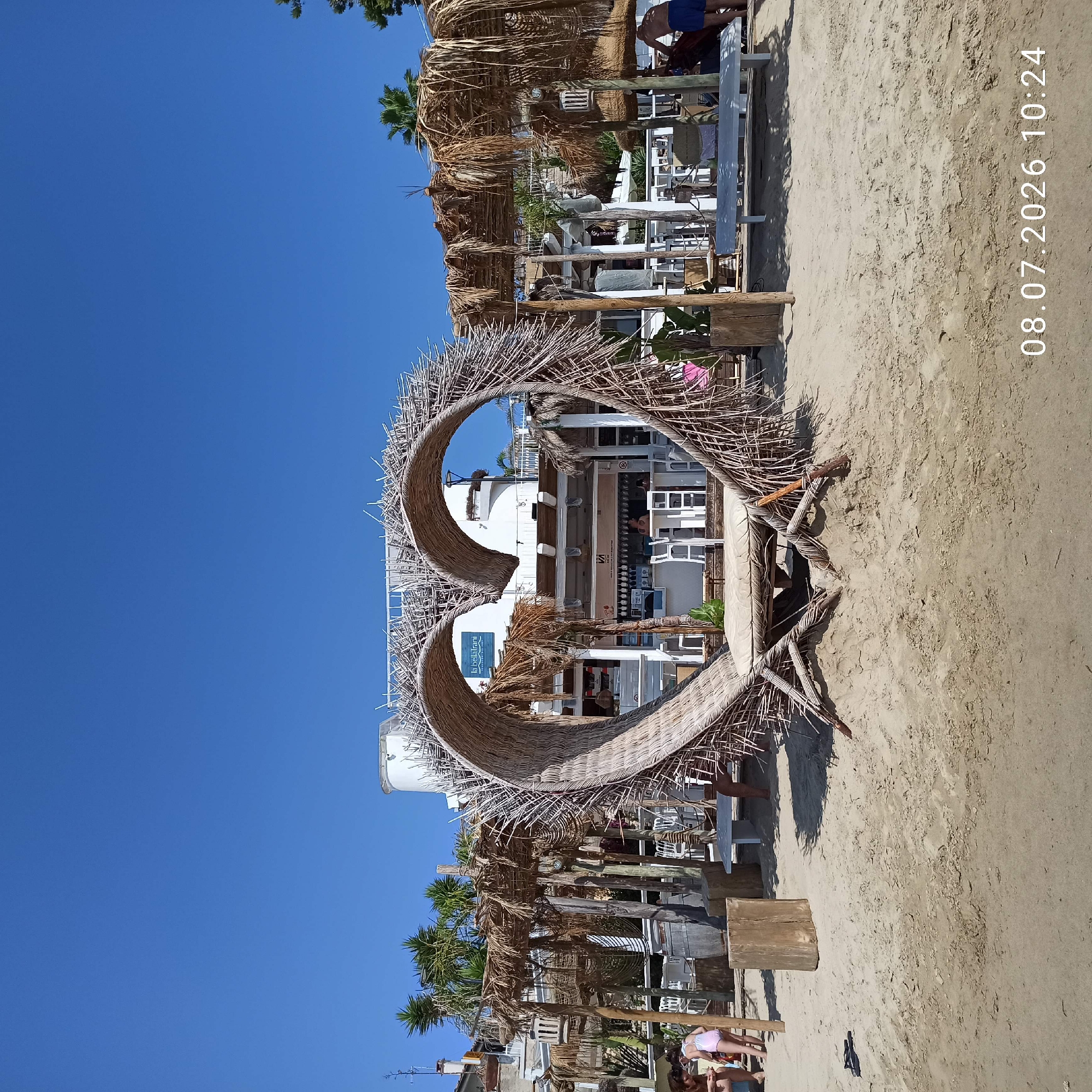The image depicts a beach scene in Trani, Italy, on a clear, sunny day. In the foreground, a large, heart-shaped structure made of woven natural materials, likely straw or reeds, dominates the center of the frame. The heart serves as an entrance to a beachside establishment, with a thatched roof canopy extending behind it.
Behind the heart, a white building with multiple levels and balconies is visible. It appears to be a restaurant or bar, with tables and chairs set up both inside and on the outdoor seating areas. A prominent sign with the text "la bellatrani" and "PARADISE BEACH" is mounted on the building's facade.
The beach itself is sandy, with some scattered debris and footprints. To the left of the heart structure, two young girls are visible, one wearing a pink swimsuit, sitting on a bench or step. Further to the left, a wooden block serves as a makeshift seat or decorative element.
To the right of the heart, more thatched canopies provide shade, with benches and tables set up underneath. A man wearing blue shorts is standing near a red object, facing away from the camera. Palm trees and other vegetation are interspersed throughout the scene, adding to the tropical beach ambiance.
The overall impression is one of a vibrant and inviting beach resort, likely a popular spot for relaxation and recreation. The timestamp "08.07.2026 10:24" is superimposed on the bottom right corner of the image.