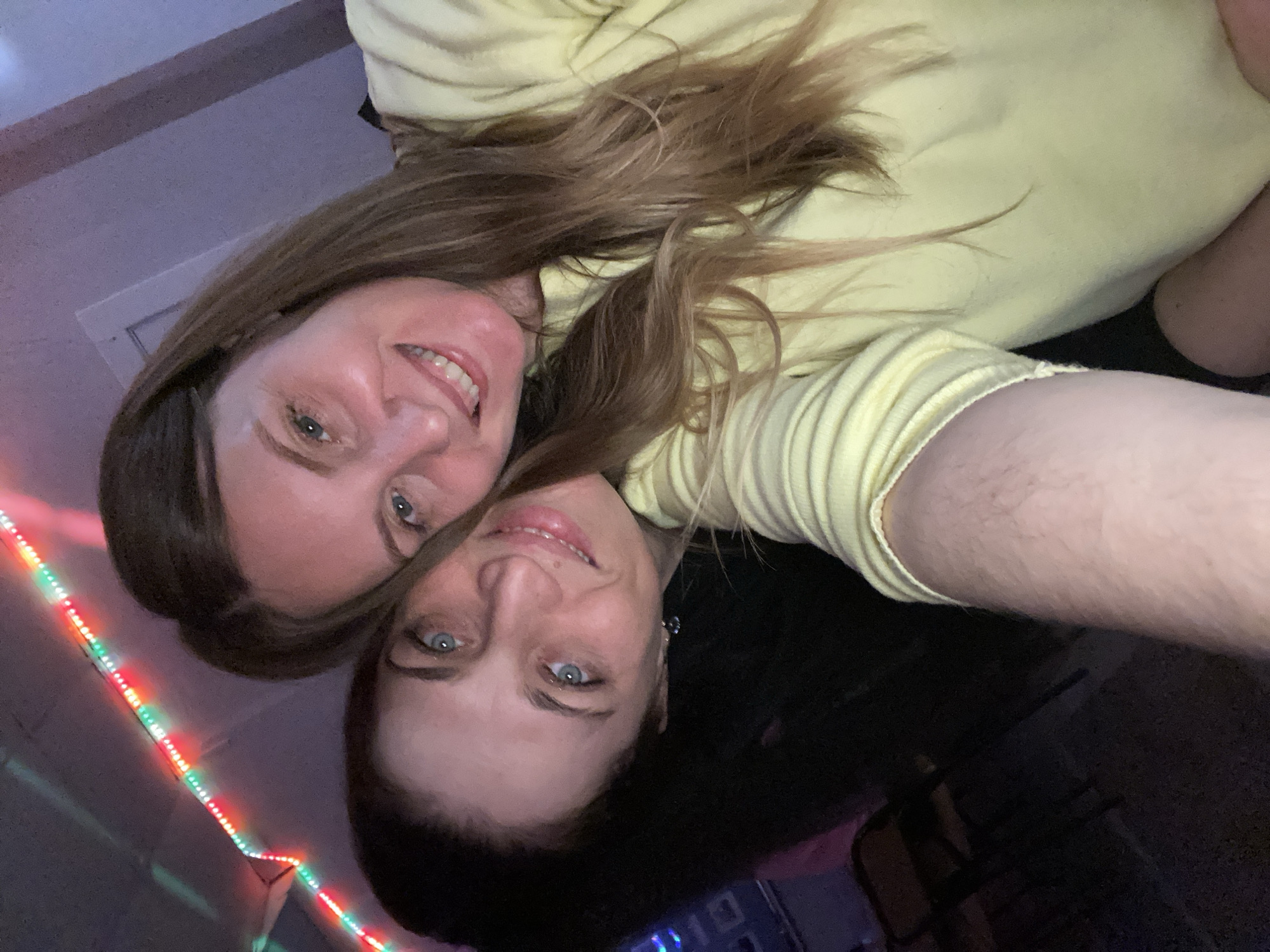 This image captures two smiling women in a close-up shot, likely a selfie taken from an overhead perspective, and is oriented ninety degrees counter-clockwise. Both women have light brown hair and blue eyes, and their faces are positioned intimately together, expressing happiness. The woman in the upper left has a broad smile, while the woman in the lower center, whose face is partially obscured by the first, also has a pleasant smile.
Both subjects are wearing light yellow, long-sleeved tops, one of which features a ribbed texture. The setting appears to be indoors with dim lighting, possibly at night. A string of colorful LED lights (red, green, and blue) runs vertically along the left side of the frame, providing a festive glow. In the top left corner, a white ceiling and a vent are visible. The overall mood is cheerful and intimate. There is no visible text in the image. The photo's location context is Noviy, Russia.