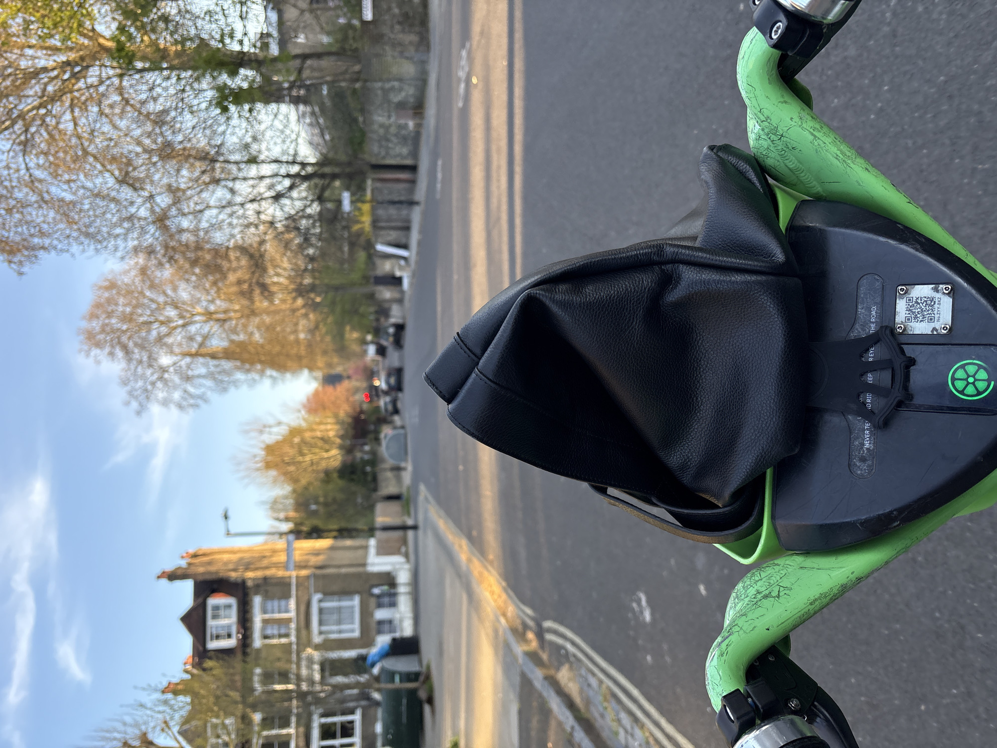 The image is a first-person perspective shot from the handlebars of a Lime scooter on a London street. The focus is on the black leather bag hanging from the handlebars, partially obscuring the view of the road ahead. The Lime scooter has green-painted handlebars and a black central panel with a Lime logo and a QR code for identification. The scooter is parked on a paved surface with a yellow line.
In the background, there is a residential street with houses and trees lining both sides. The buildings are a mix of brick and stucco, with some having visible windows and architectural details. The sky is blue with some clouds, and the light suggests it is either morning or late afternoon. The road is visible in the distance, with some cars. The photo appears to have been taken in a residential area of London, United Kingdom, likely a quieter street with trees and well-maintained houses.
