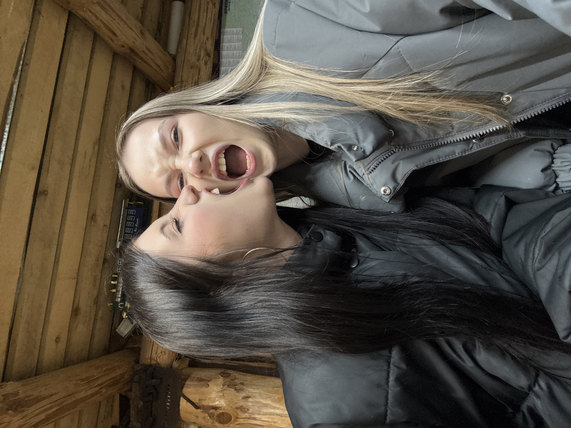 Two women are posing closely together. One woman with blonde hair has her mouth wide open. The second woman with dark hair has her mouth slightly open. Both women are wearing dark puffer jackets. The photo appears to be taken indoors. The immediate environment is a wooden structure. In the background, there's a device with small boxes labeled in Russian. It is difficult to pinpoint the exact location in Фоки, Russia.