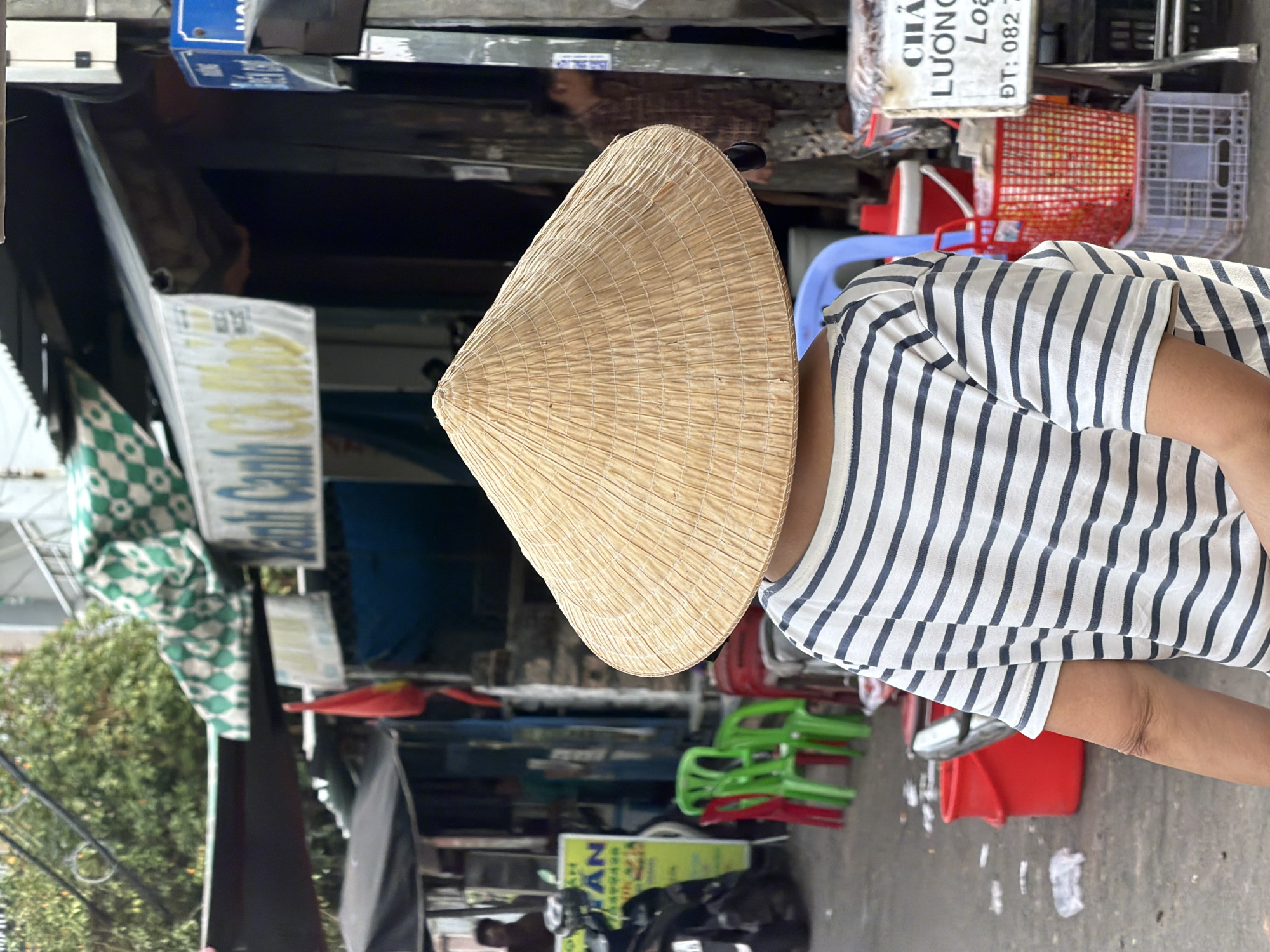 In Nha Trang, Vietnam, a person wearing a conical hat and a striped t-shirt is photographed from behind, facing away from the camera. They stand on a street with a paved surface, where some litter is visible. In the background, there are market stalls with awnings and a variety of objects, including red and green plastic chairs, buckets, and baskets. A scooter is parked to the left, and a sign with Vietnamese text is visible on a stall. The overall scene suggests a bustling outdoor market environment.