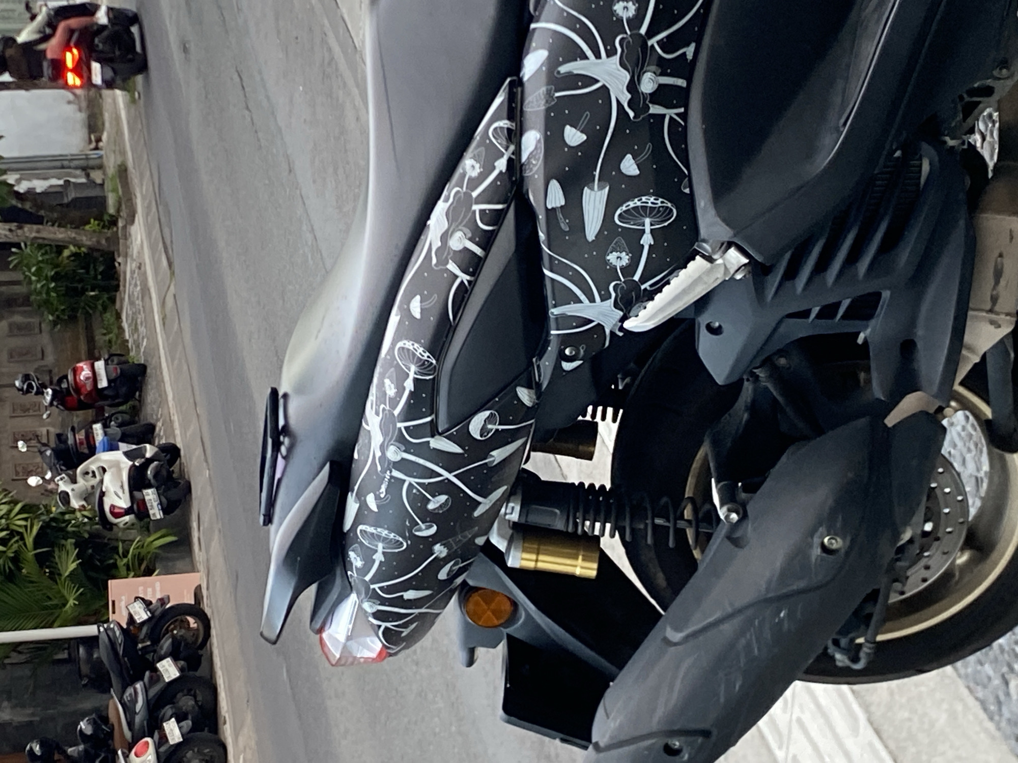 The image shows a close-up view of the rear section of a black motorcycle parked on a street in Mengwi, Indonesia. The motorcycle features a decorative decal with a white pattern of mushrooms and celestial elements on its body panels. In the background, other motorcycles are parked along the side of the street, some with riders. A sidewalk and a road are visible, with some greenery and buildings in the further background. The lighting suggests it is daytime.